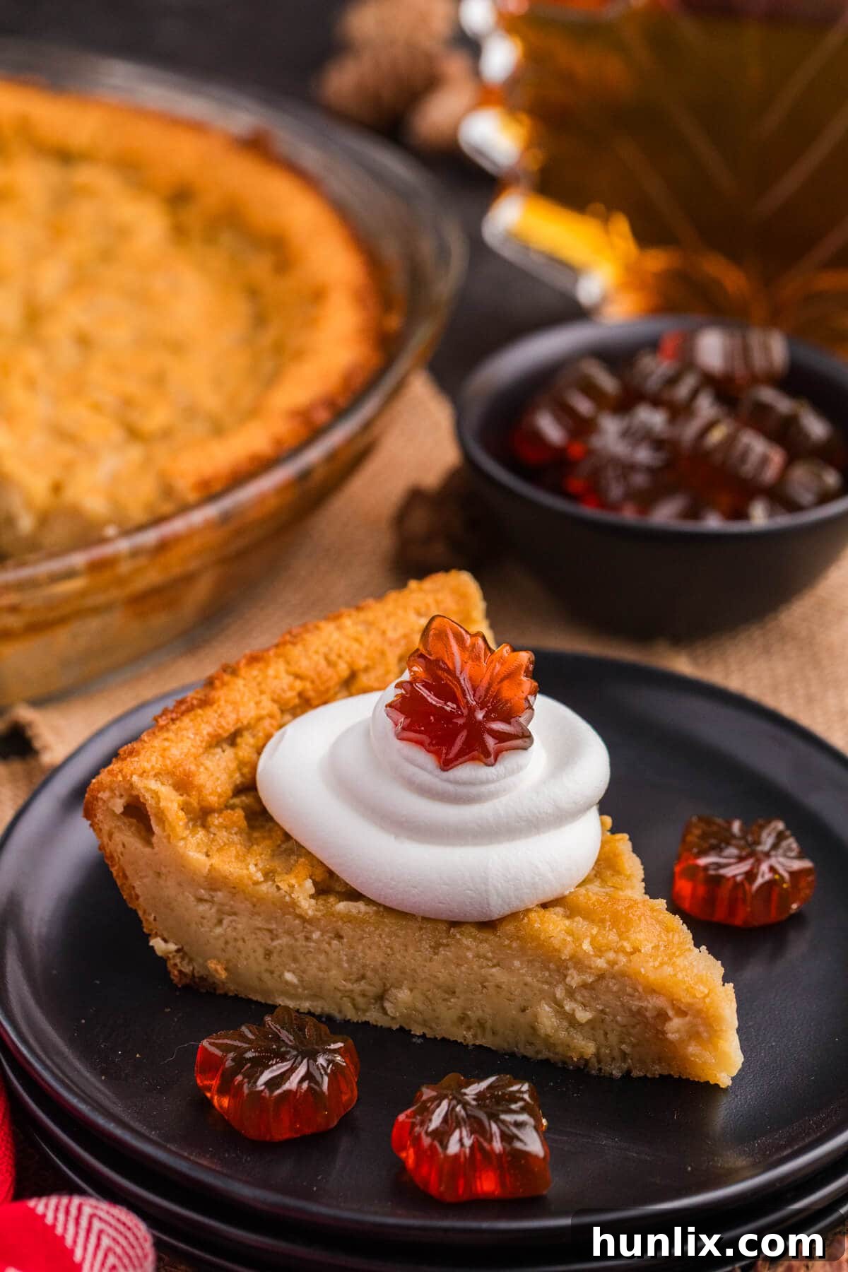 Maple Magic Pie 2 A perfectly golden slice of Maple Impossible Pie, topped with a dollop of whipped cream and a delicate maple leaf candy, sits on a white plate with a fork beside it.
