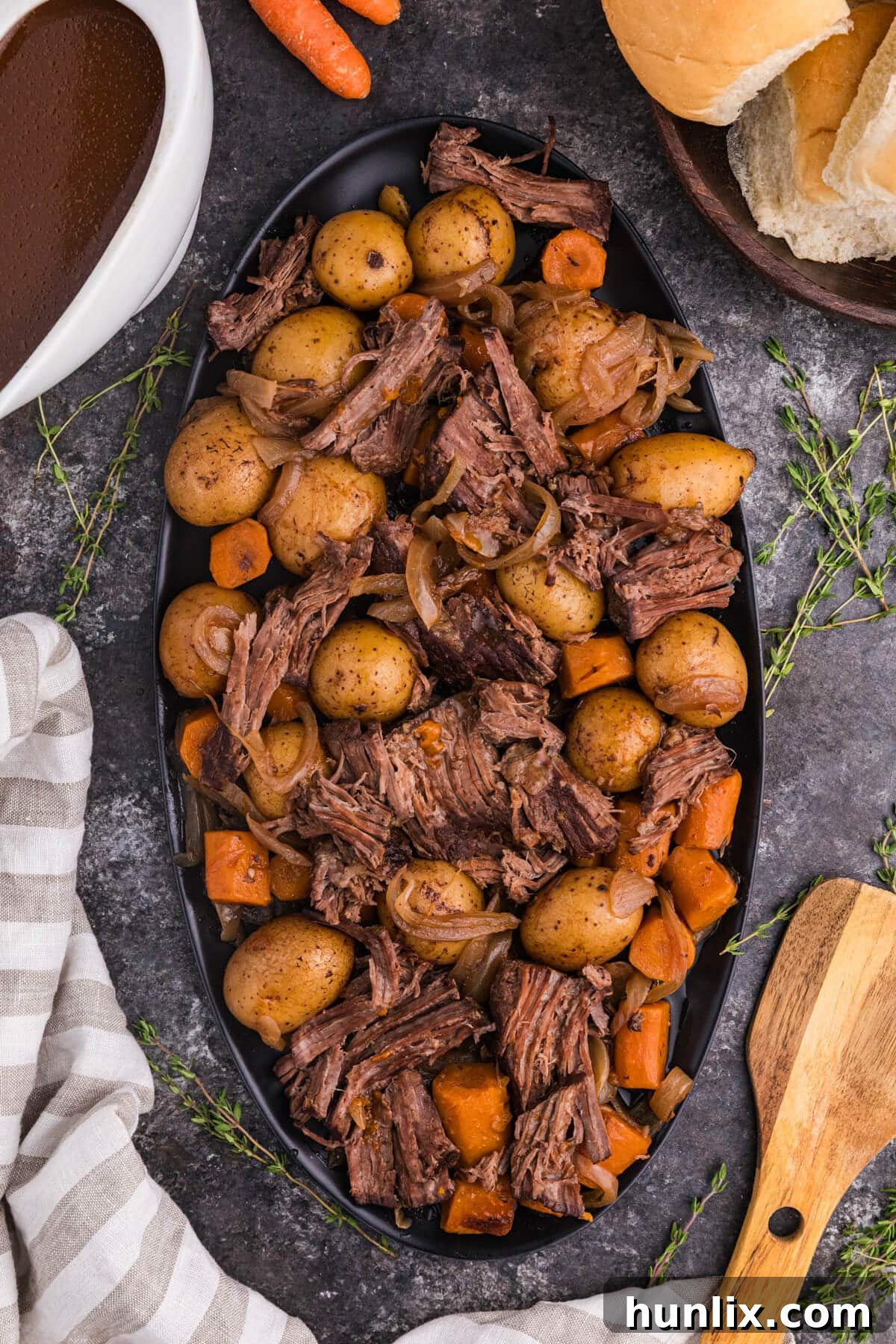 Fall Apart Slow Cooker Pot Roast 2 A serving platter filled with tender shredded slow cooker pot roast, baby potatoes, carrots, and onions, surrounded by a bowl of rich brown gravy, fresh thyme sprigs, dinner rolls, and a wooden spoon on a dark surface.