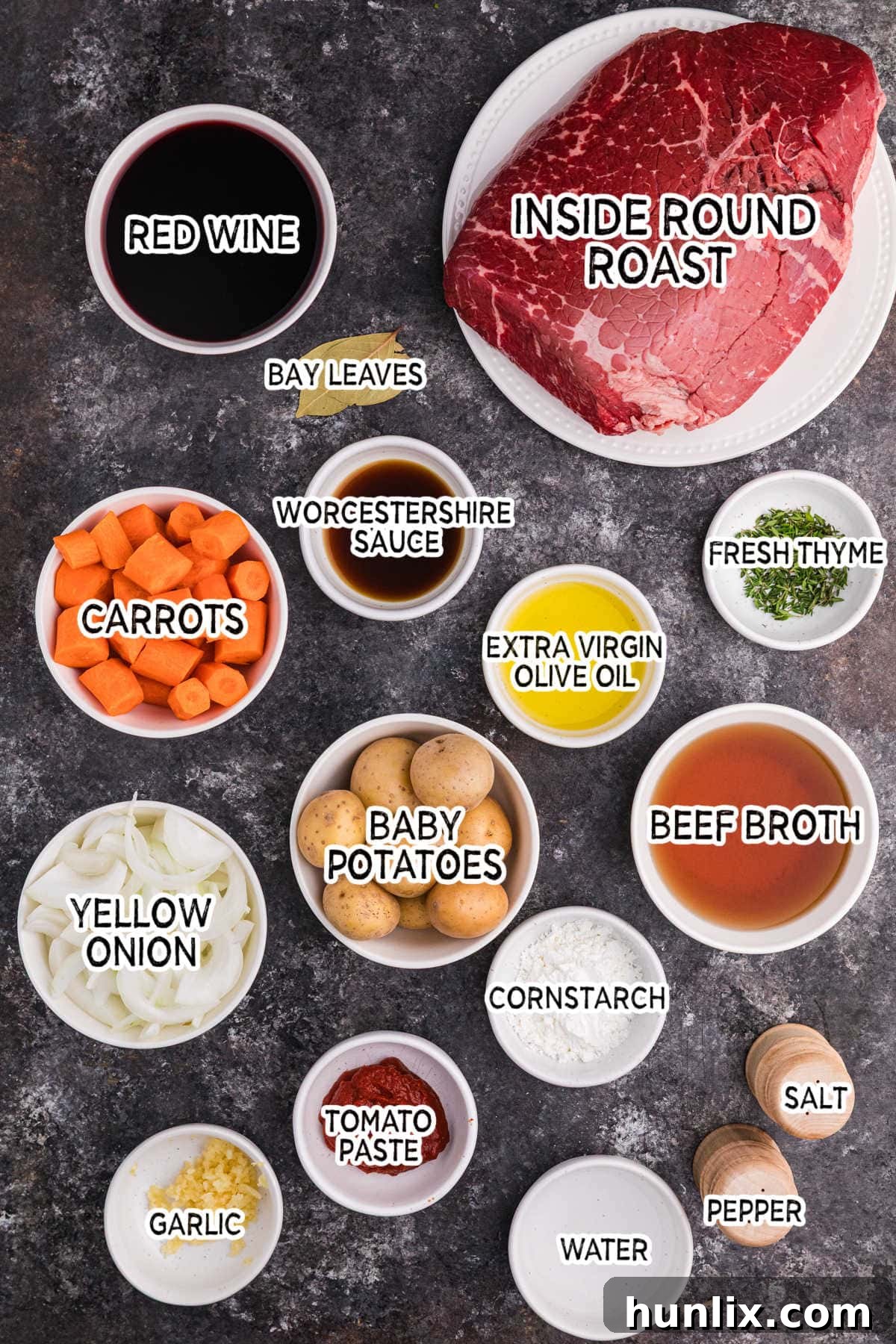 Fall Apart Slow Cooker Pot Roast 3 Overhead view of labeled ingredients including beef roast, baby potatoes, carrots, onion, garlic, tomato paste, beef broth, red wine, Worcestershire sauce, thyme, bay leaves, olive oil, cornstarch, water, salt, and pepper on a dark surface.