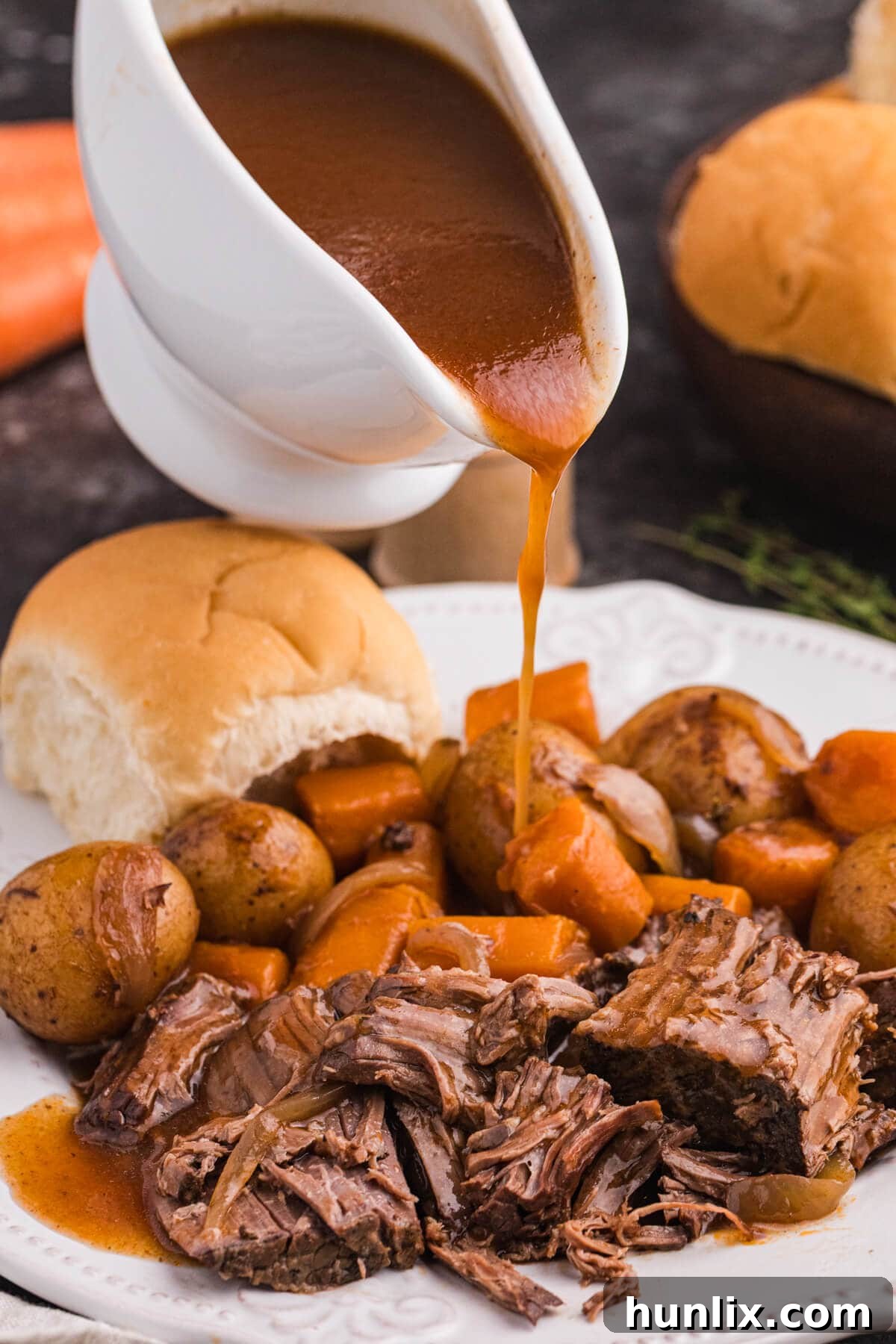 Fall Apart Slow Cooker Pot Roast 8 Gravy being poured from a white gravy boat over tender beef, carrots, and baby potatoes on a plate with a dinner roll in the background.