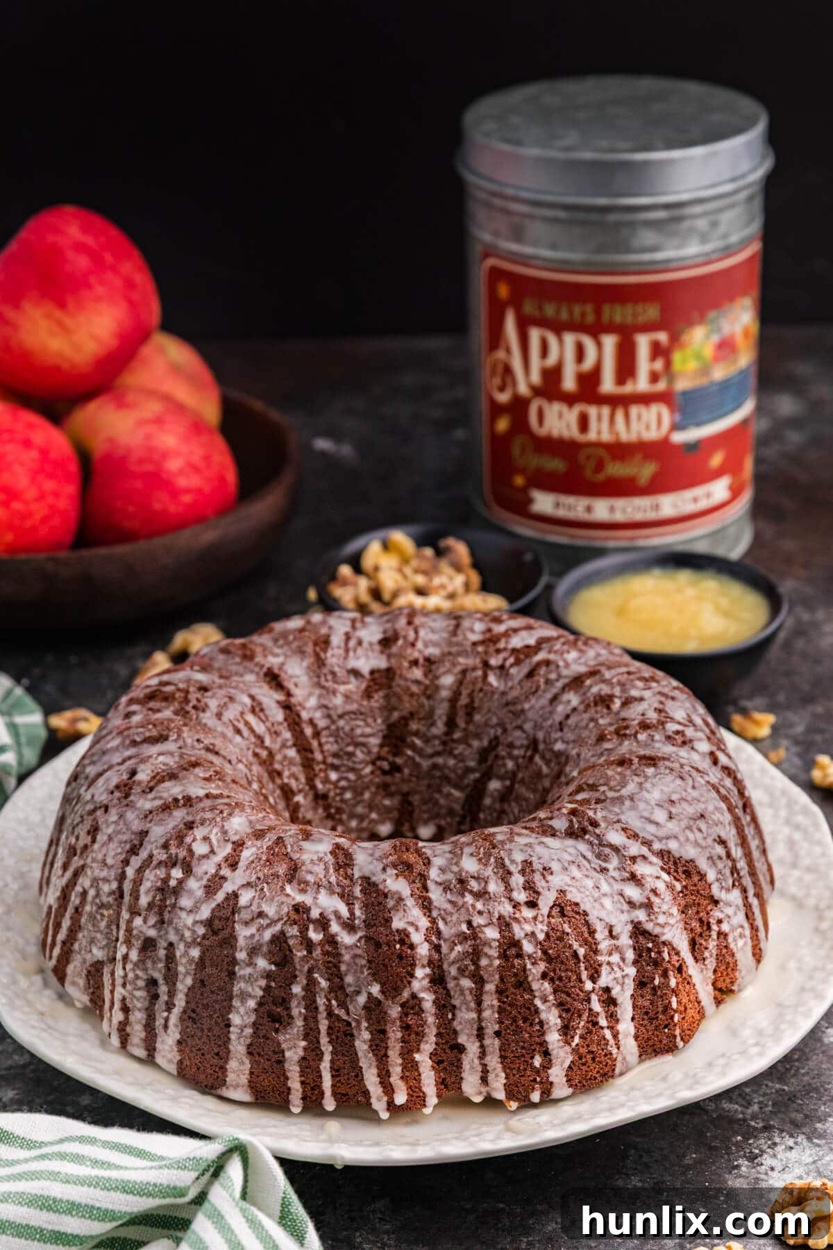 A beautifully glazed applesauce cake sits on a white plate, drizzled with a thin vanilla glaze. Fresh red apples, a rustic tin, a small dish of applesauce, and scattered walnuts are arranged in the background on a dark surface, evoking a cozy, autumnal feel.