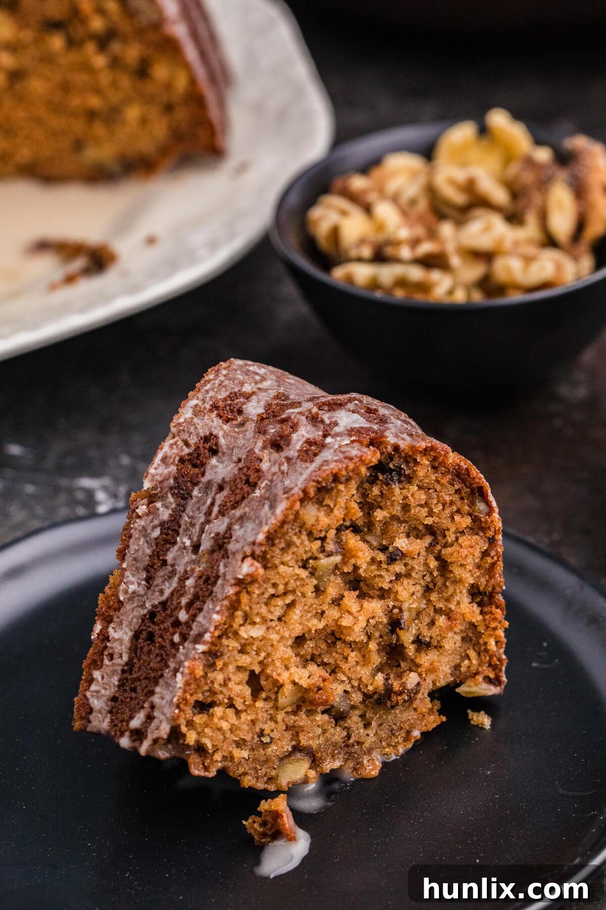 A single, perfectly cut slice of applesauce cake sits on a dark plate, showcasing its moist, spiced crumb and visible walnut pieces, topped with a light glaze. The remaining bundt cake and a small bowl of walnuts are softly blurred in the background.