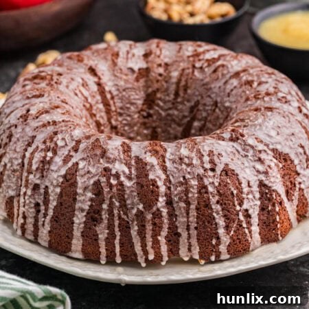 A full applesauce cake, elegantly covered in a thin vanilla glaze, rests on a pristine white plate. In the blurred background, fresh red apples, chopped walnuts, and a small bowl of applesauce are visible, alongside a green-striped cloth, creating an inviting culinary scene.