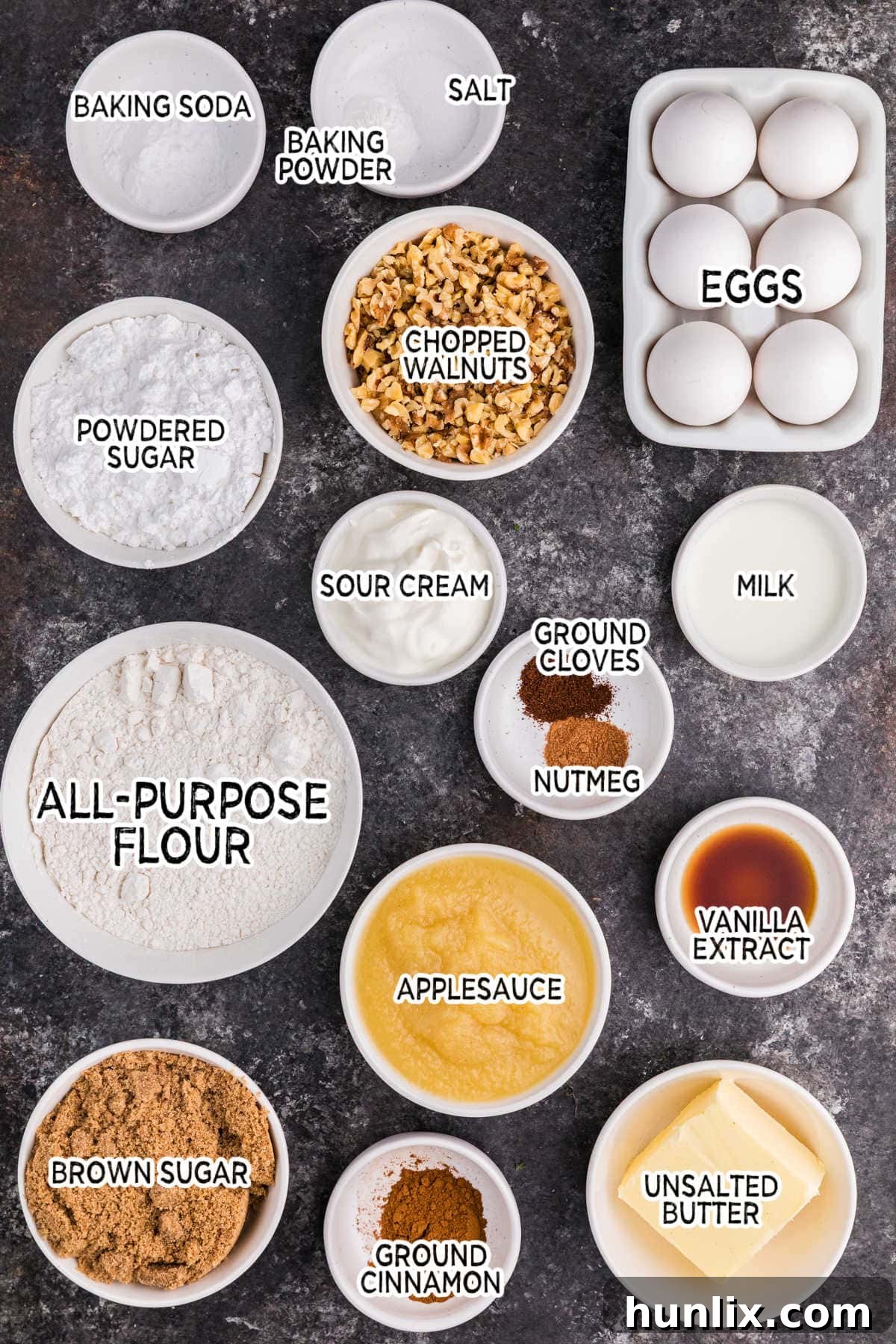 All the dry and wet ingredients for applesauce cake are neatly arranged on a dark background, showcasing baking essentials.