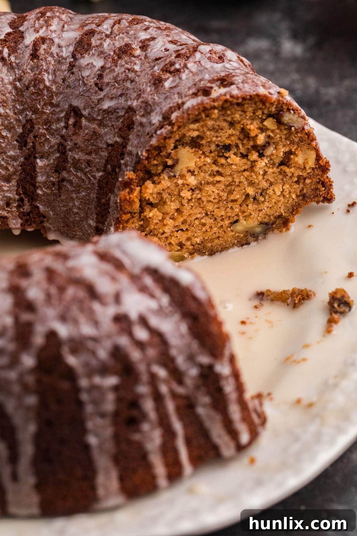 A close-up view of a perfectly sliced bundt cake, revealing its moist, spiced interior studded with small walnut pieces. A delicate vanilla glaze drips down the sides onto the plate, inviting a bite.