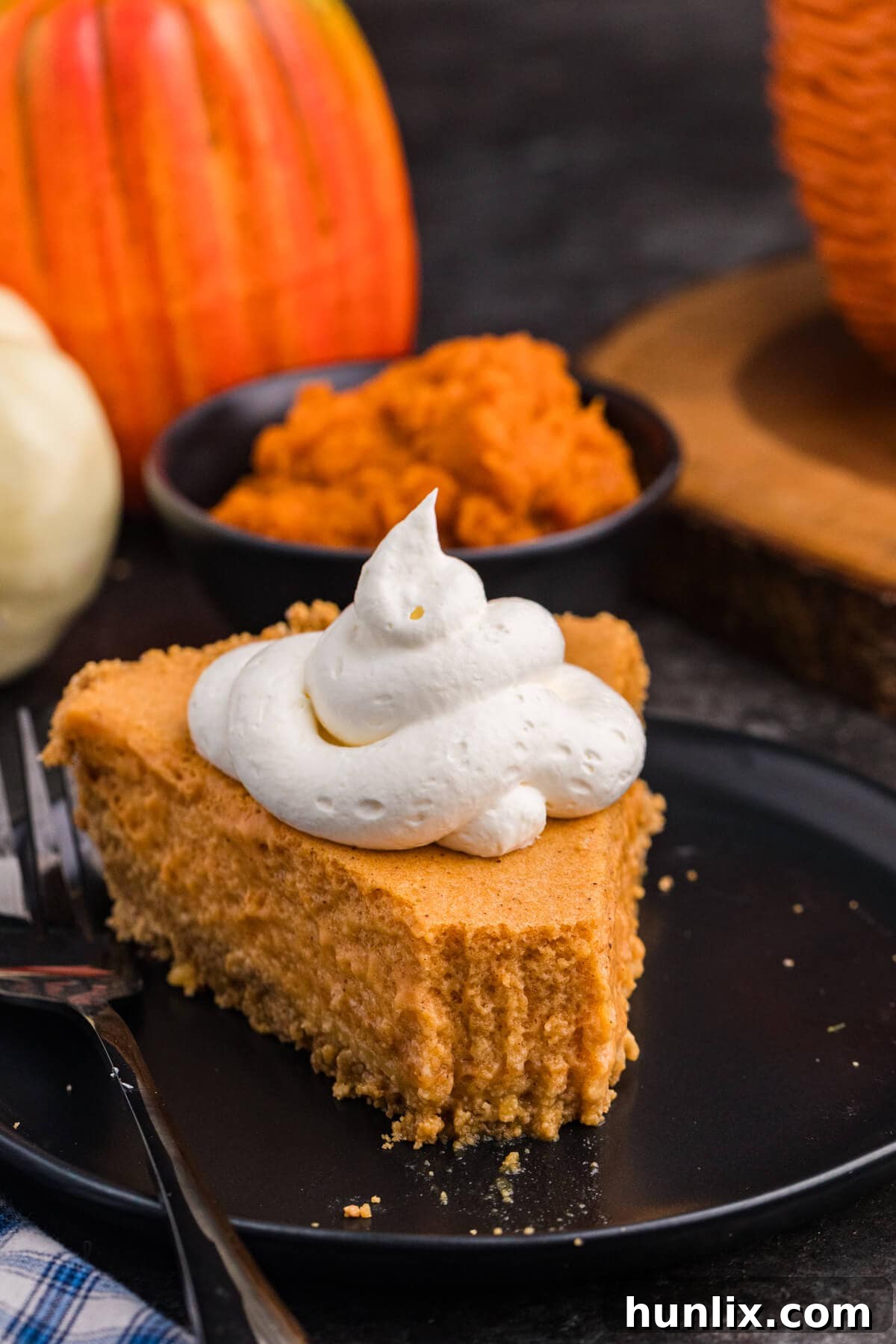 Whipped Pumpkin Delight 10 A slice of light, creamy pumpkin pie topped with a swirl of whipped cream sits on a black plate, with pumpkins and a bowl of pumpkin purée blurred in the background.