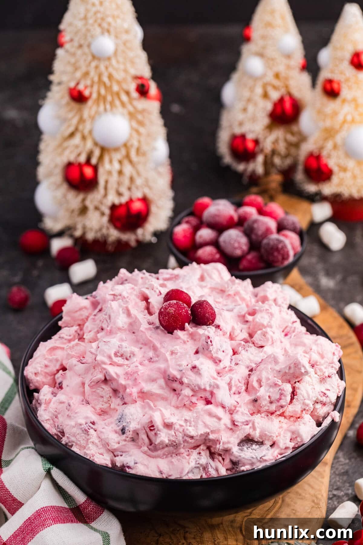 A beautifully presented Cranberry fluff salad in a black bowl, garnished with fresh cranberries, surrounded by mini marshmallows and elegant Christmas decorations featuring festive trees and frosted berries, evoking a cozy holiday atmosphere.