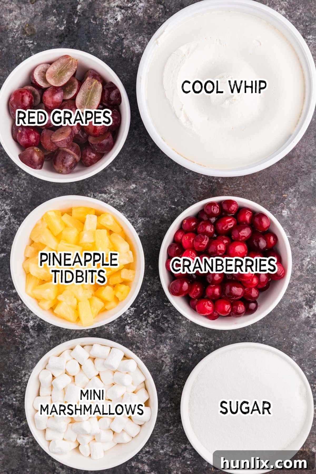 A collection of fresh and pantry ingredients laid out on a dark background, including vibrant red cranberries, a bowl of sugar, red grapes, canned pineapple, mini marshmallows, and a container of Cool Whip, ready for preparing Cranberry Fluff Salad.