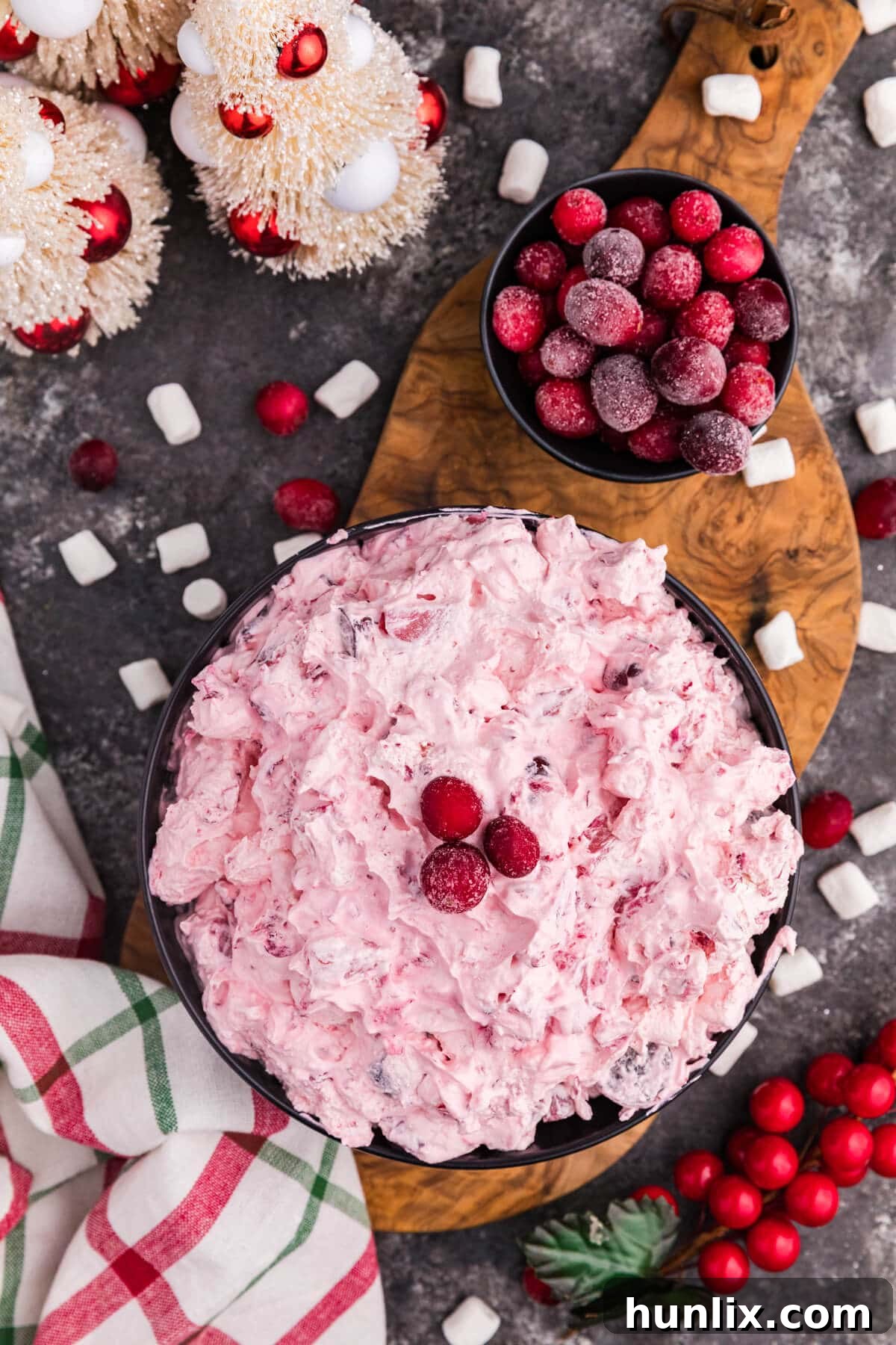A vibrant pink whipped dessert topped with sugared cranberries in a black bowl, surrounded by scattered mini marshmallows, frosted berries, holiday decorations, and a red and green plaid napkin on a rustic dark surface, ready to be served.