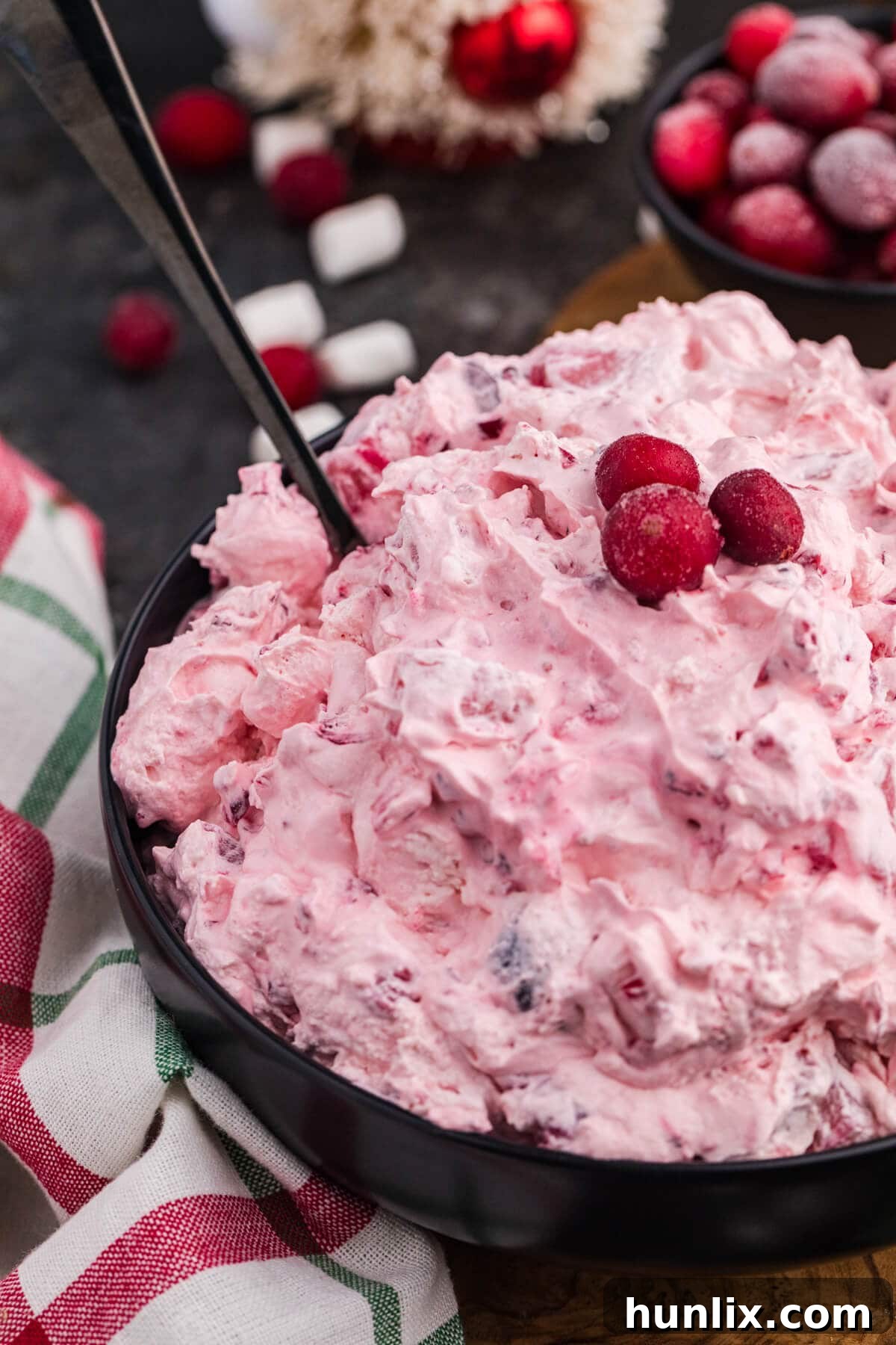 A close-up shot of a fluffy pink holiday dessert in a black bowl, elegantly garnished with sugared cranberries, with a serving spoon resting beside it. The scene includes a red and green plaid napkin, festive decorations, and scattered mini marshmallows in the background, highlighting its delicious texture.