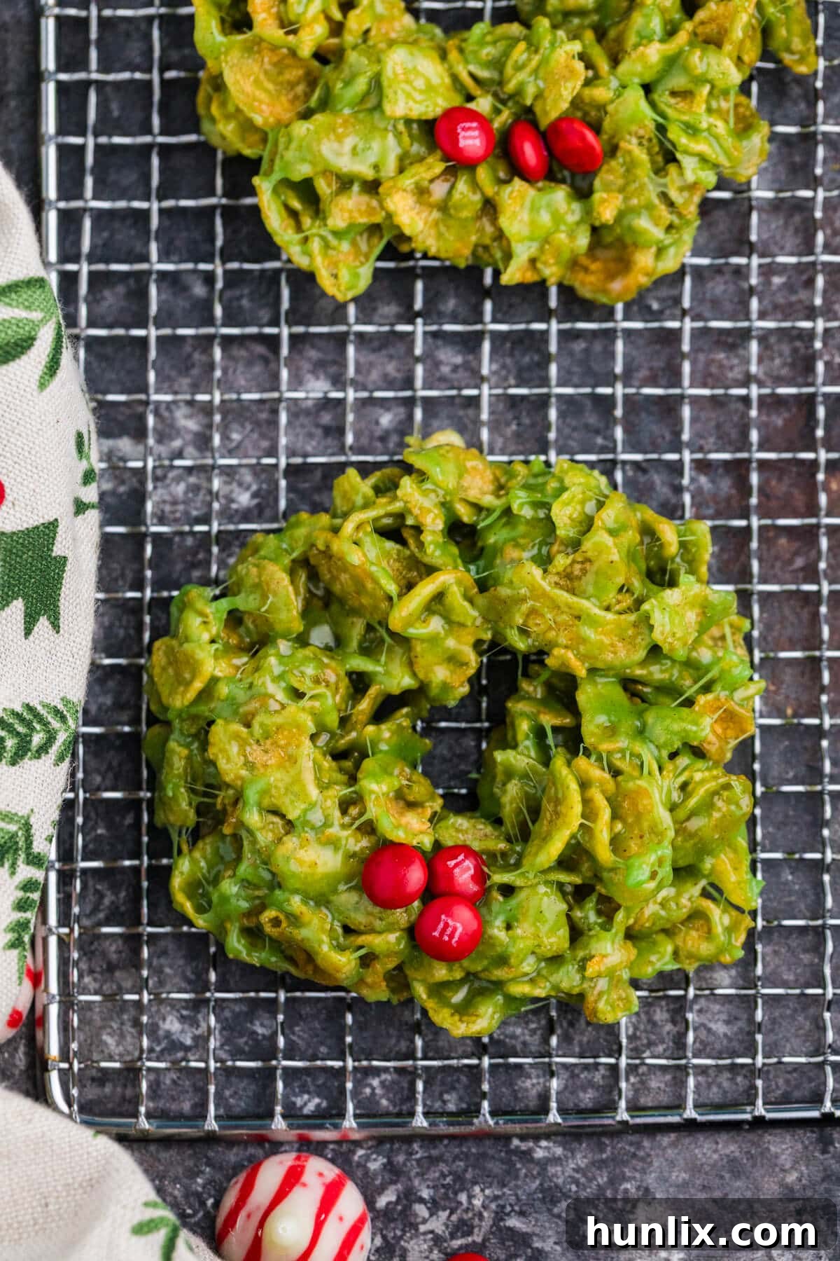 Crispy Cereal Wreaths 6 Green marshmallow-coated cornflake wreath cookies cooling on a wire rack, each decorated with red M&M candies to resemble holly berries, set against a dark stone background with festive fabric and a peppermint candy nearby.