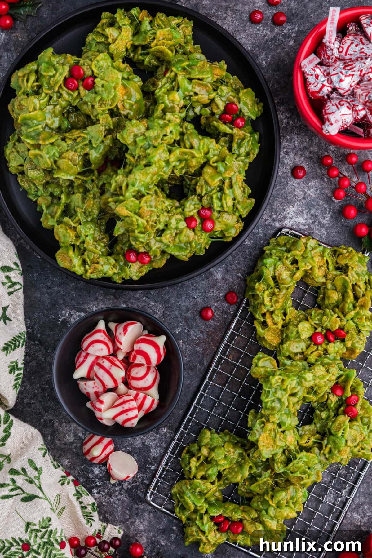 Crispy Cereal Wreaths 7 Top-down view of green cornflake wreath cookies decorated with red candies, displayed on a black plate and cooling rack. Surrounding elements include a bowl of striped peppermint chocolates, scattered red candy pieces, and festive greenery on a holiday-themed cloth.