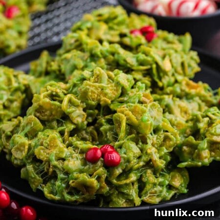 Close-up of green cornflake wreath cookies on a black plate, with red M&M candies as holly berries. Background details include peppermint swirl chocolates in a bowl and festive red berry decorations.