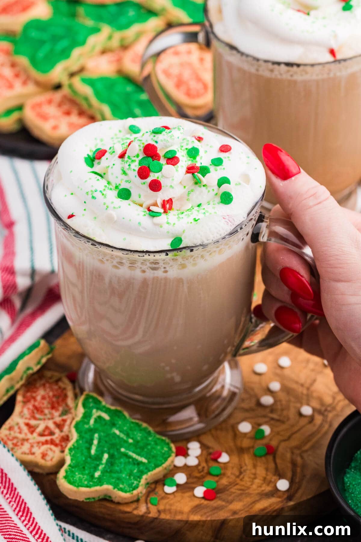 Close-up of a hand with red nail polish holding a festive glass mug filled with latte topped with whipped cream and red, green, and white holiday sprinkles, surrounded by decorated Christmas cookies and colorful sugar sprinkles on a wooden board.
