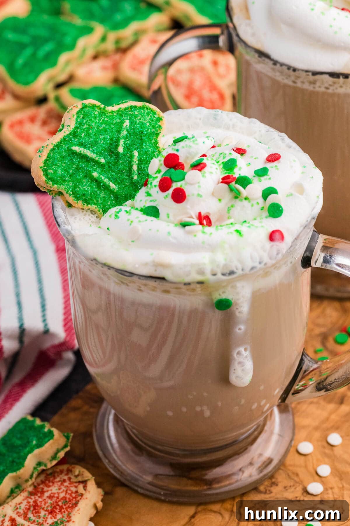 Glass mug filled with a frothy holiday latte, topped with whipped cream, festive red, green, and white sprinkles, and a green sugar-coated Christmas tree cookie resting on top, with more decorated cookies and a striped towel in the background.
