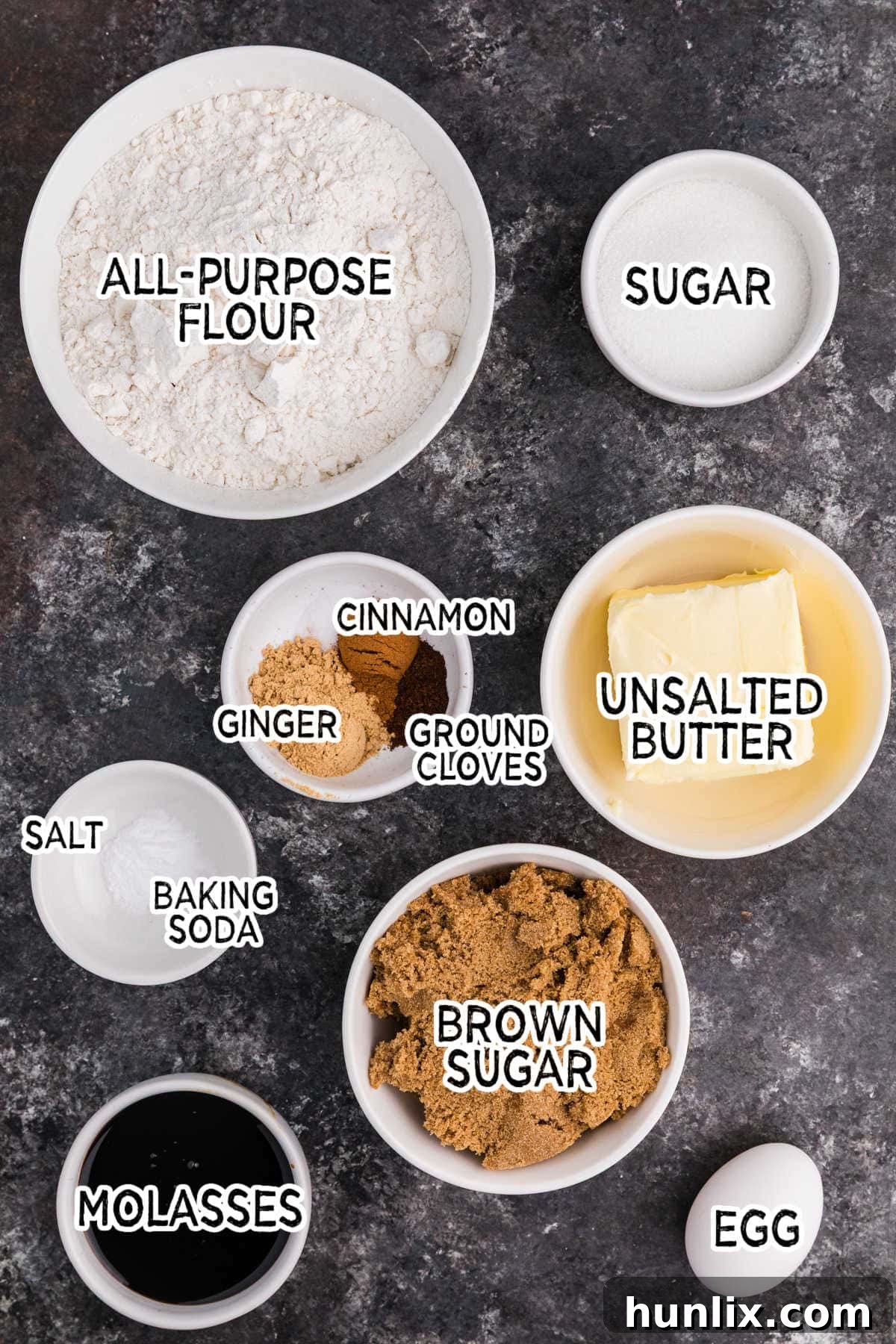 A collection of ingredients for ginger molasses cookies arranged on a black background, including softened butter, brown sugar, eggs, molasses, flour, and various spices.