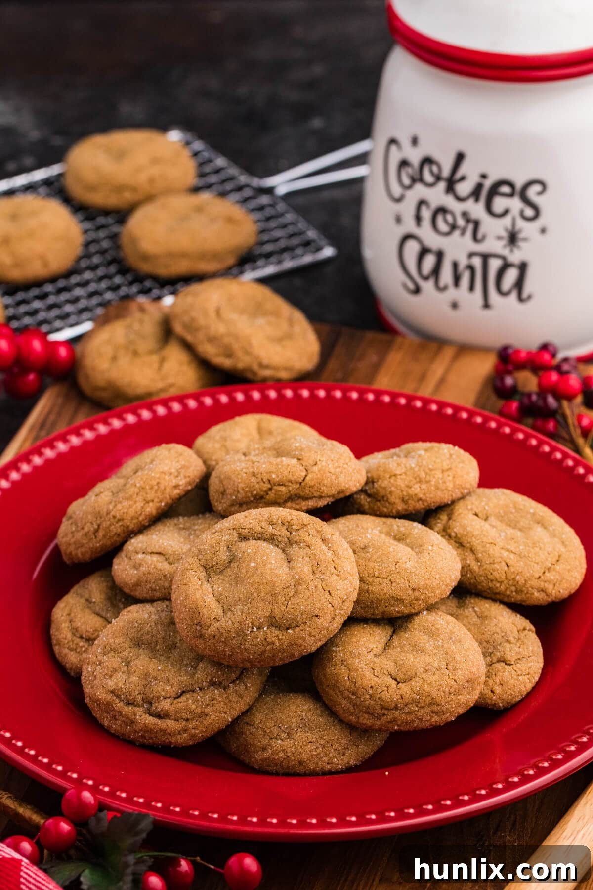 A festive red plate piled with soft, sugar-dusted holiday cookies, surrounded by red berries, a cooling rack with more cookies, and a white “Cookies for Santa” jar in the background.