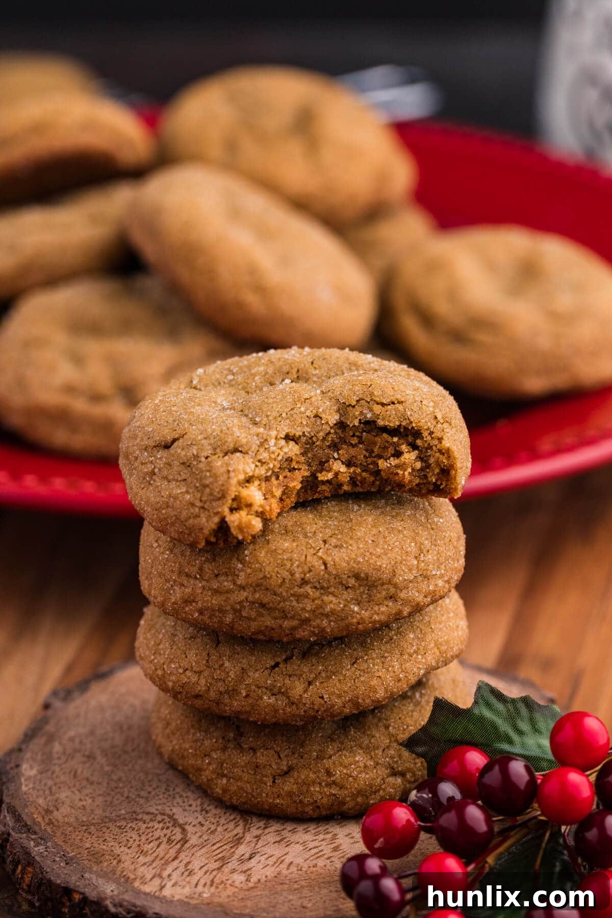 A stack of soft, sugar-coated ginger molasses cookies on a rustic wooden slice, with a bite taken out of the top cookie, surrounded by festive red berries.
