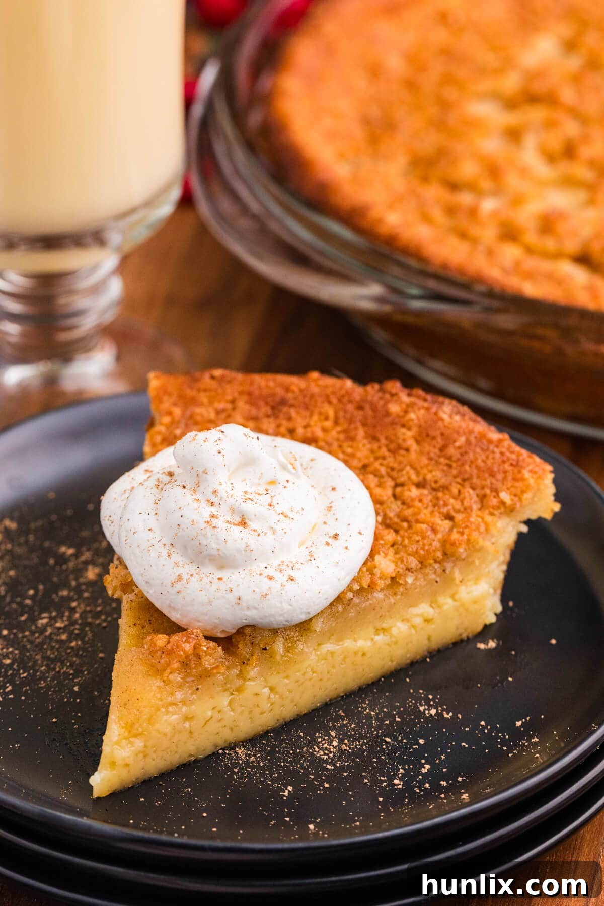 Slice of impossible eggnog pie topped with a swirl of whipped cream and a dusting of ground nutmeg, served on a black plate with the full pie and a glass of eggnog in the background; festive holiday dessert featuring a creamy custard filling and golden brown crust.