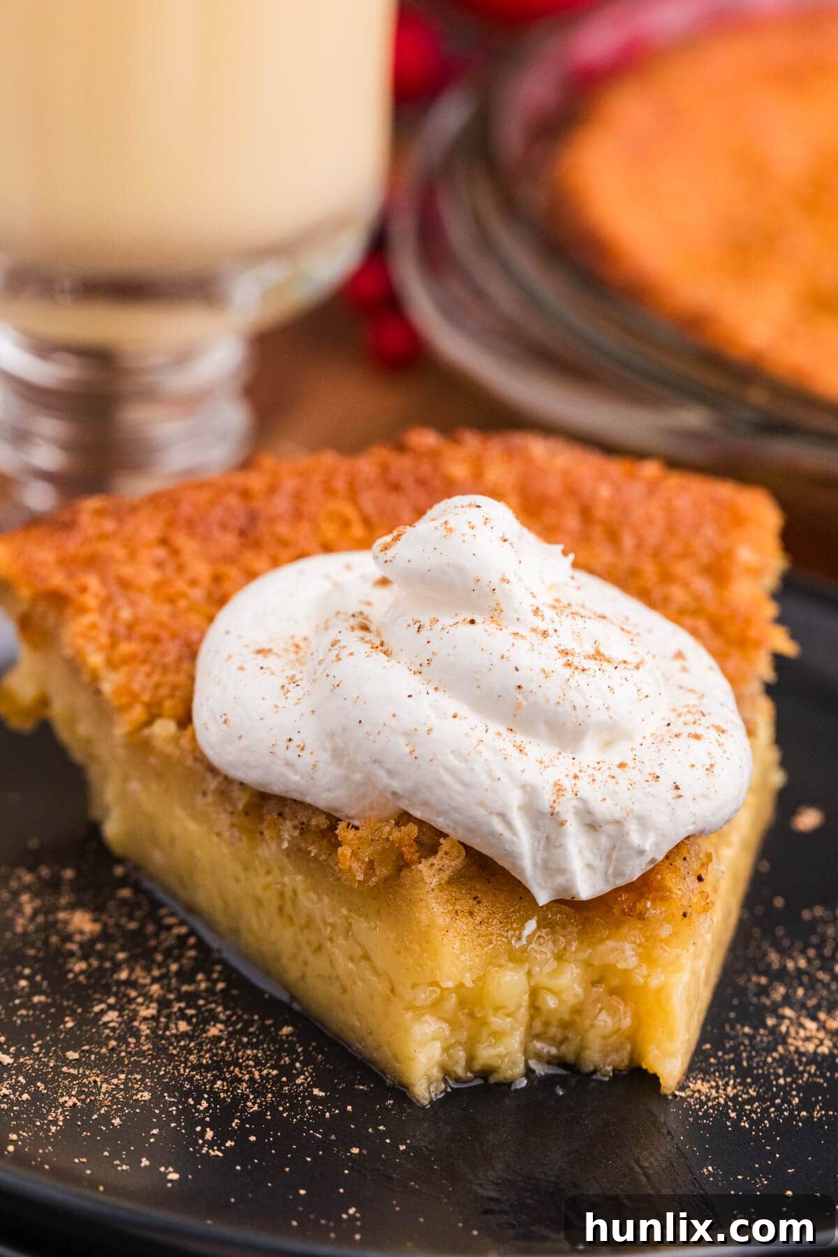Slice of creamy custard pie with golden crust, topped with whipped cream and sprinkled with nutmeg on a black plate.