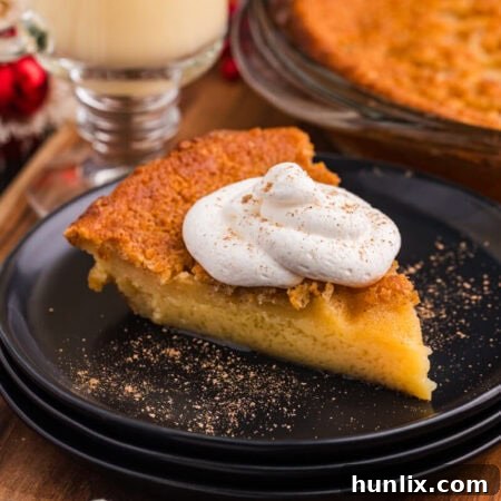 Slice of golden custard pie topped with whipped cream and nutmeg, served on a black plate with festive holiday decor and a glass of eggnog in the background.
