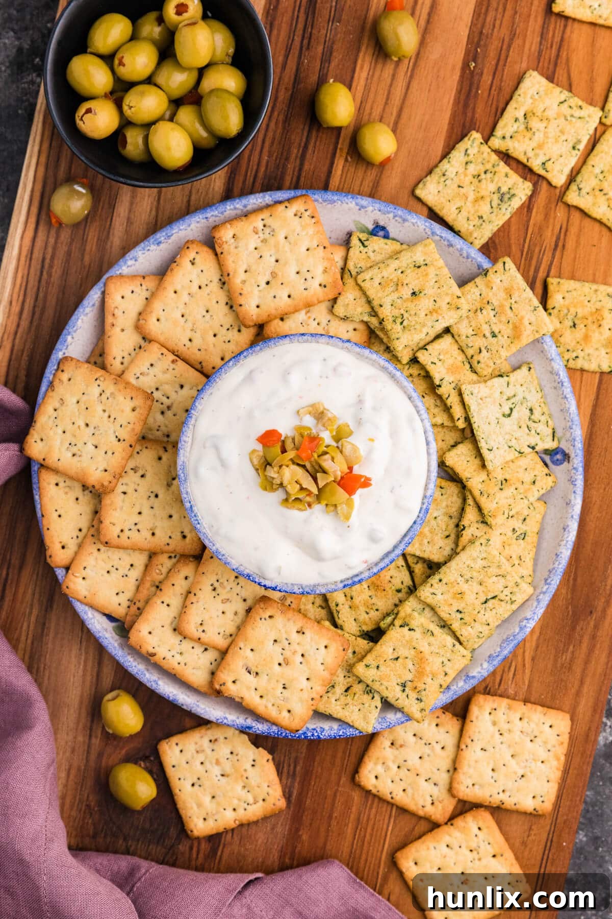 Creamy olive dip topped with finely chopped green and pimento-stuffed olives, served alongside a variety of seasoned and herb crackers on an elegant wooden platter, perfect for any gathering.
