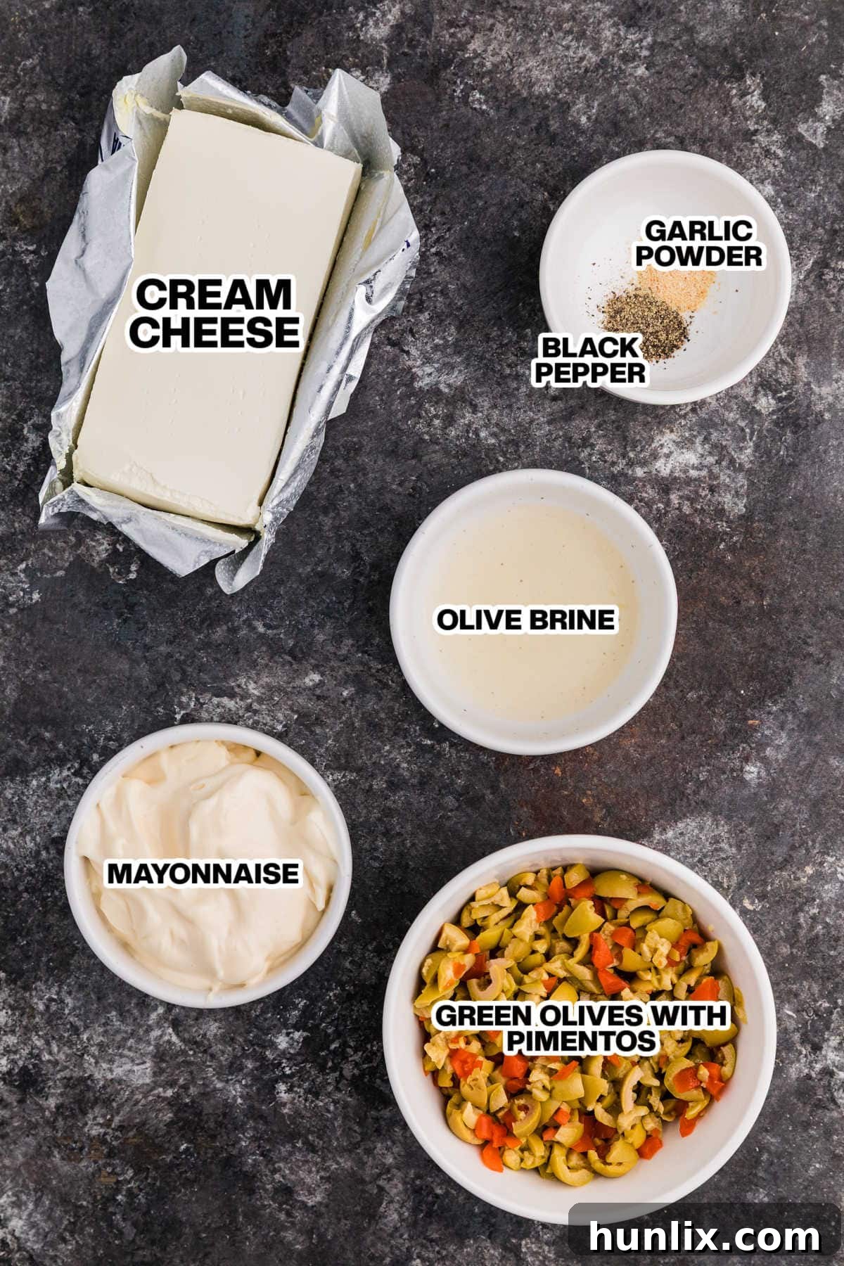 A selection of fresh ingredients laid out on a dark background, including cream cheese, mayonnaise, a jar of green olives with pimentos, olive brine, garlic powder, and black pepper, all ready for making the delicious olive dip.