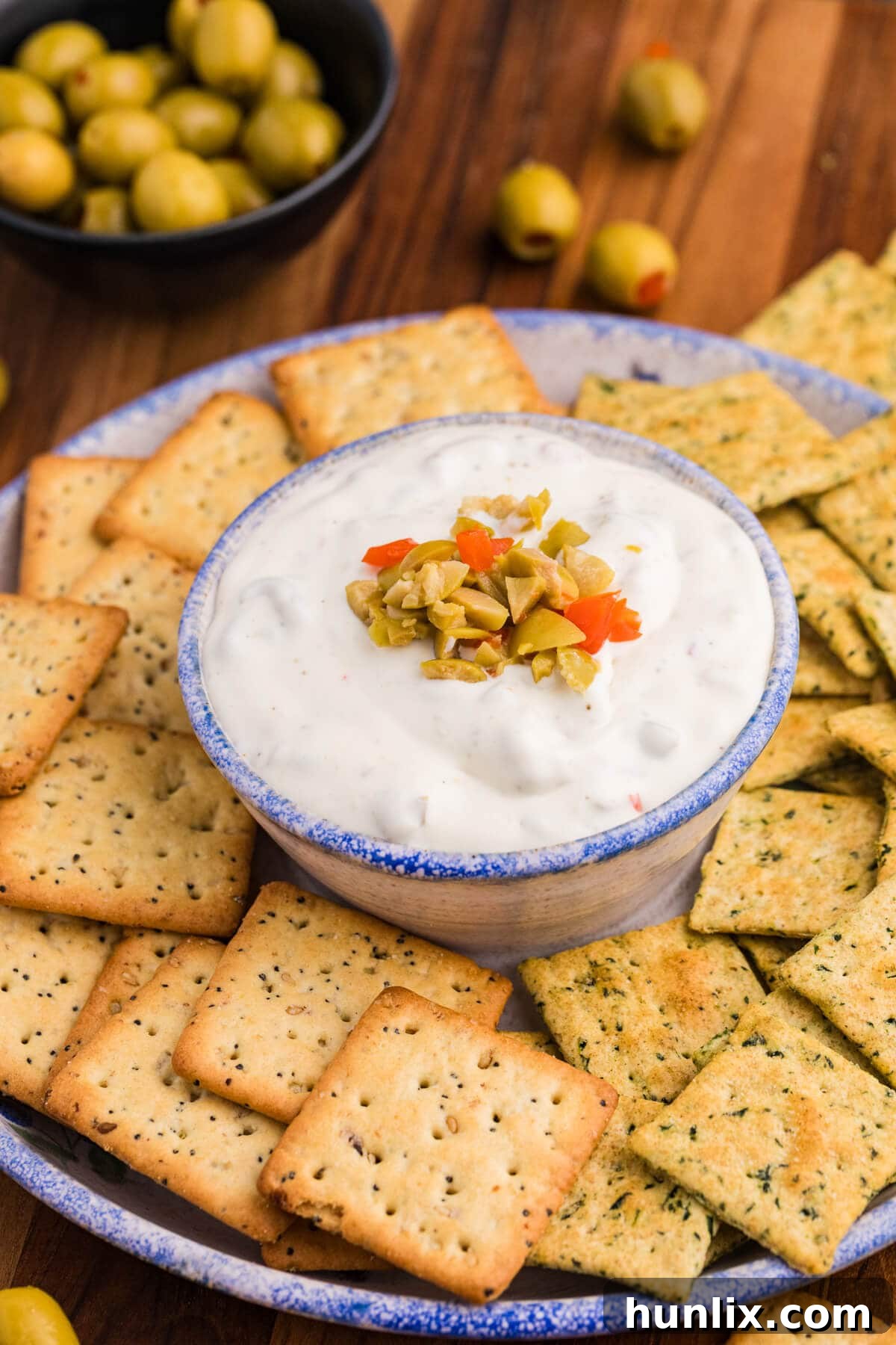 A close-up shot of a creamy white dip, generously topped with finely chopped green and vibrant red pimento-stuffed olives. The dip is surrounded by a selection of herb and multigrain square crackers on an elegant ceramic plate, showcasing its appetizing appeal.
