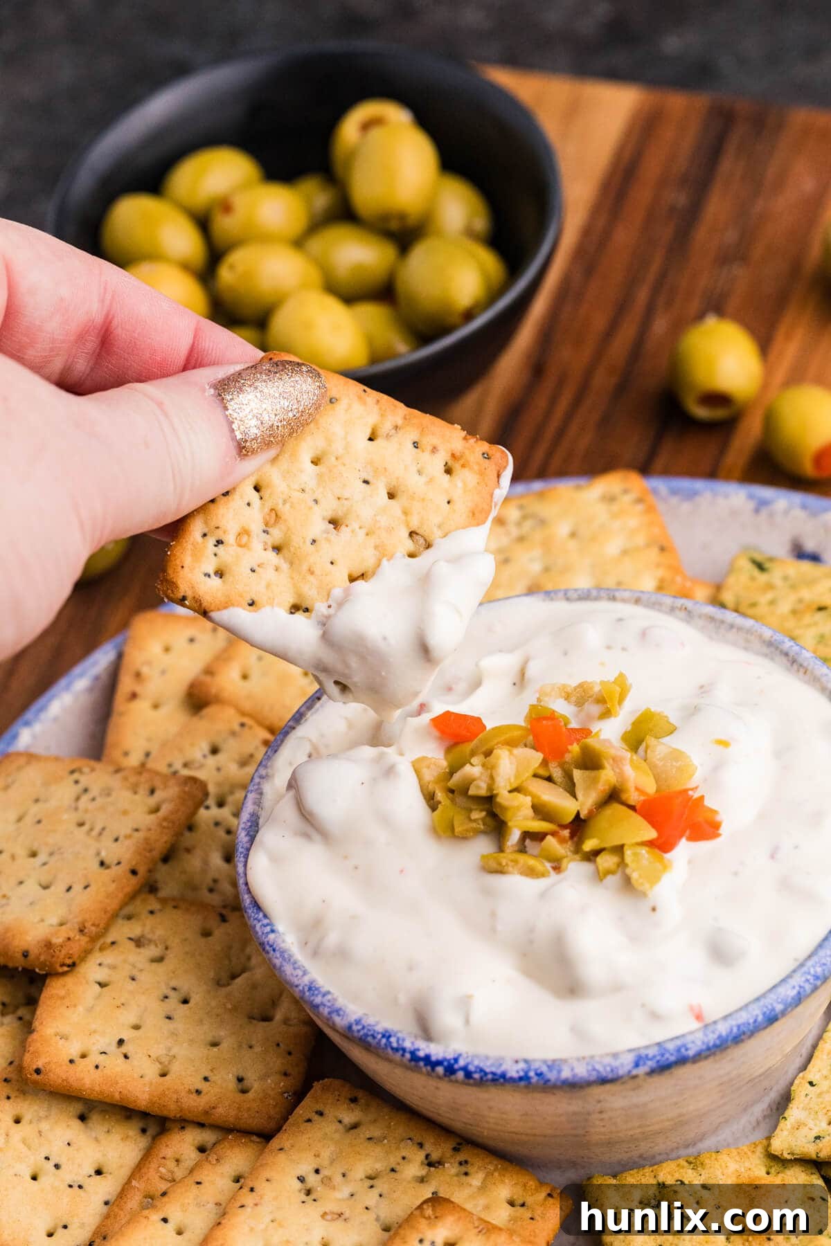 A hand gently dipping a seeded square cracker into a ceramic bowl of creamy white olive dip, which is beautifully topped with chopped green and red pimento-stuffed olives. In the background, a small bowl of whole green olives and a variety of assorted crackers are visible, highlighting a perfect serving setup.