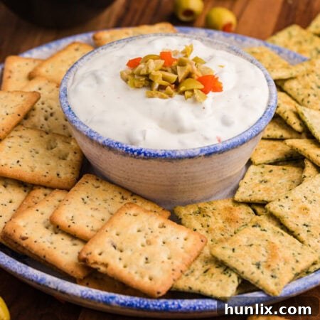 Close-up of a ceramic bowl filled with creamy white dip topped with chopped green and red pimento-stuffed olives, surrounded by seasoned multigrain and herb-flavored square crackers on a rustic serving plate.