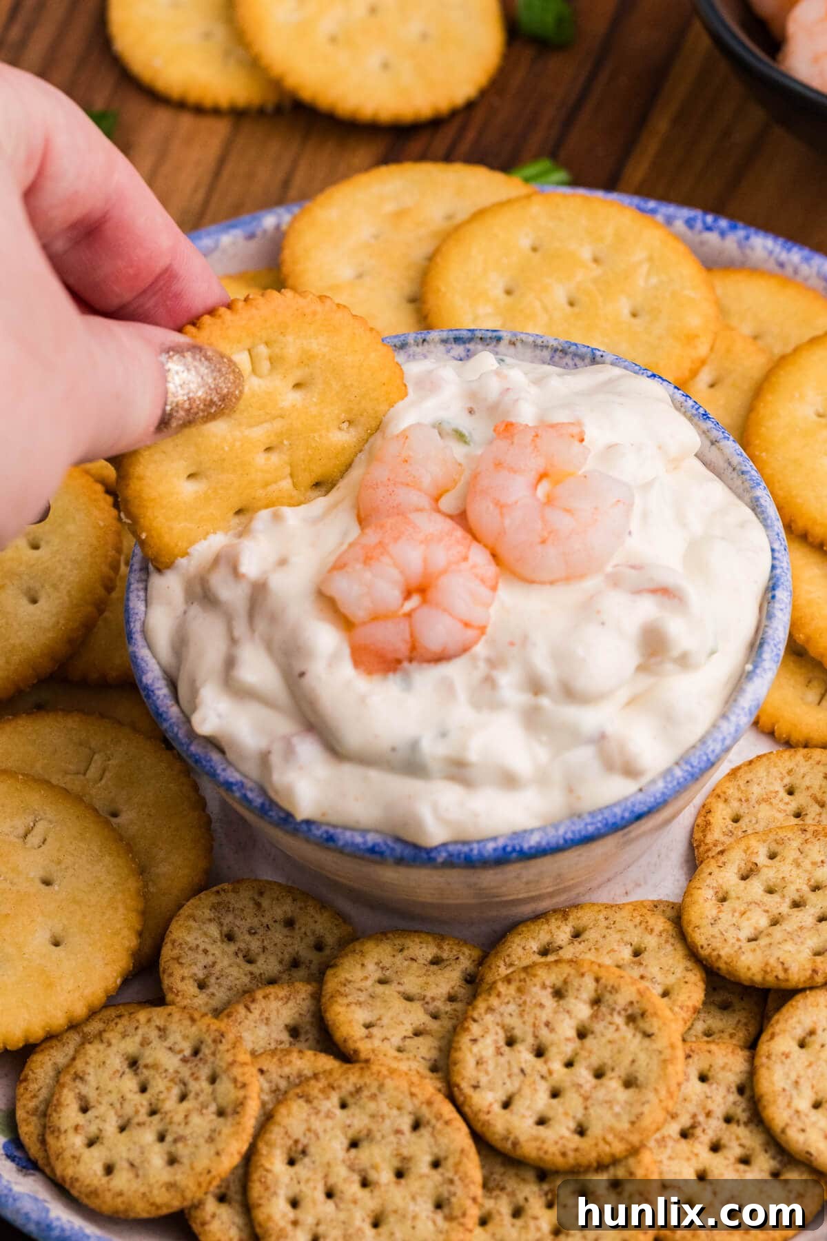 Irresistible Shrimp Dip 5 Hand dipping a Ritz cracker into shrimp-topped creamy dip, surrounded by assorted crackers.