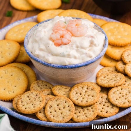 Creamy dip topped with shrimp in a ceramic bowl, served with Ritz and multigrain crackers on a rustic plate.