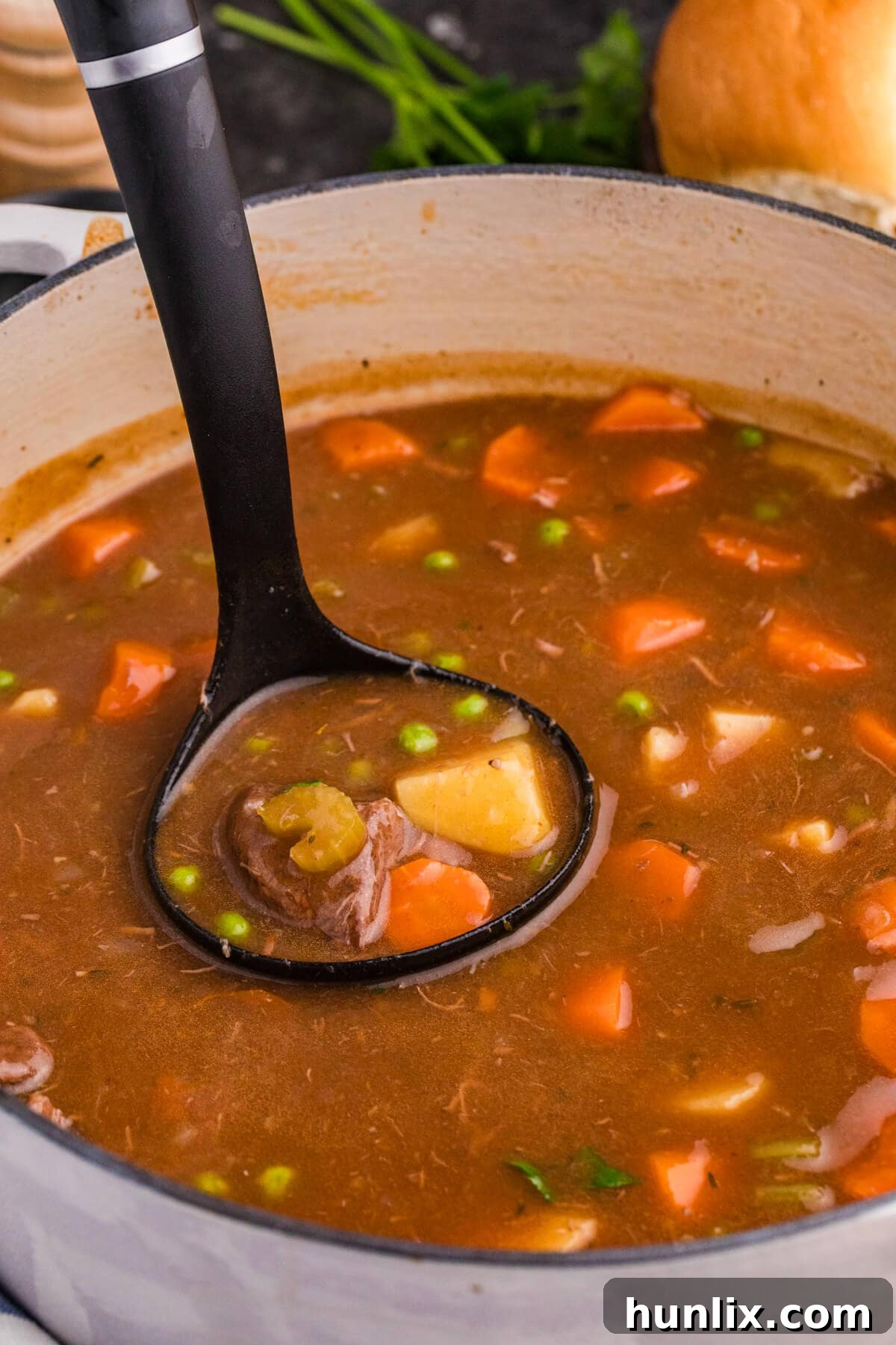 A close-up of a hearty beef stew served in a rustic Dutch oven with a ladle, featuring generous chunks of beef, tender potatoes, vibrant carrots, green peas, and celery in a rich, dark brown broth, garnished with fresh herbs.