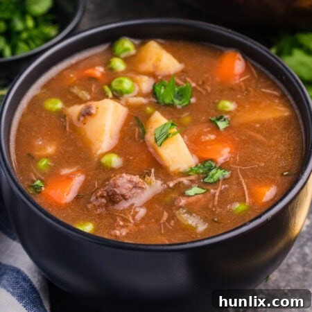 Side view of a bowl of stew with beef, carrots, potatoes, peas, and parsley, next to a towel and herbs.
