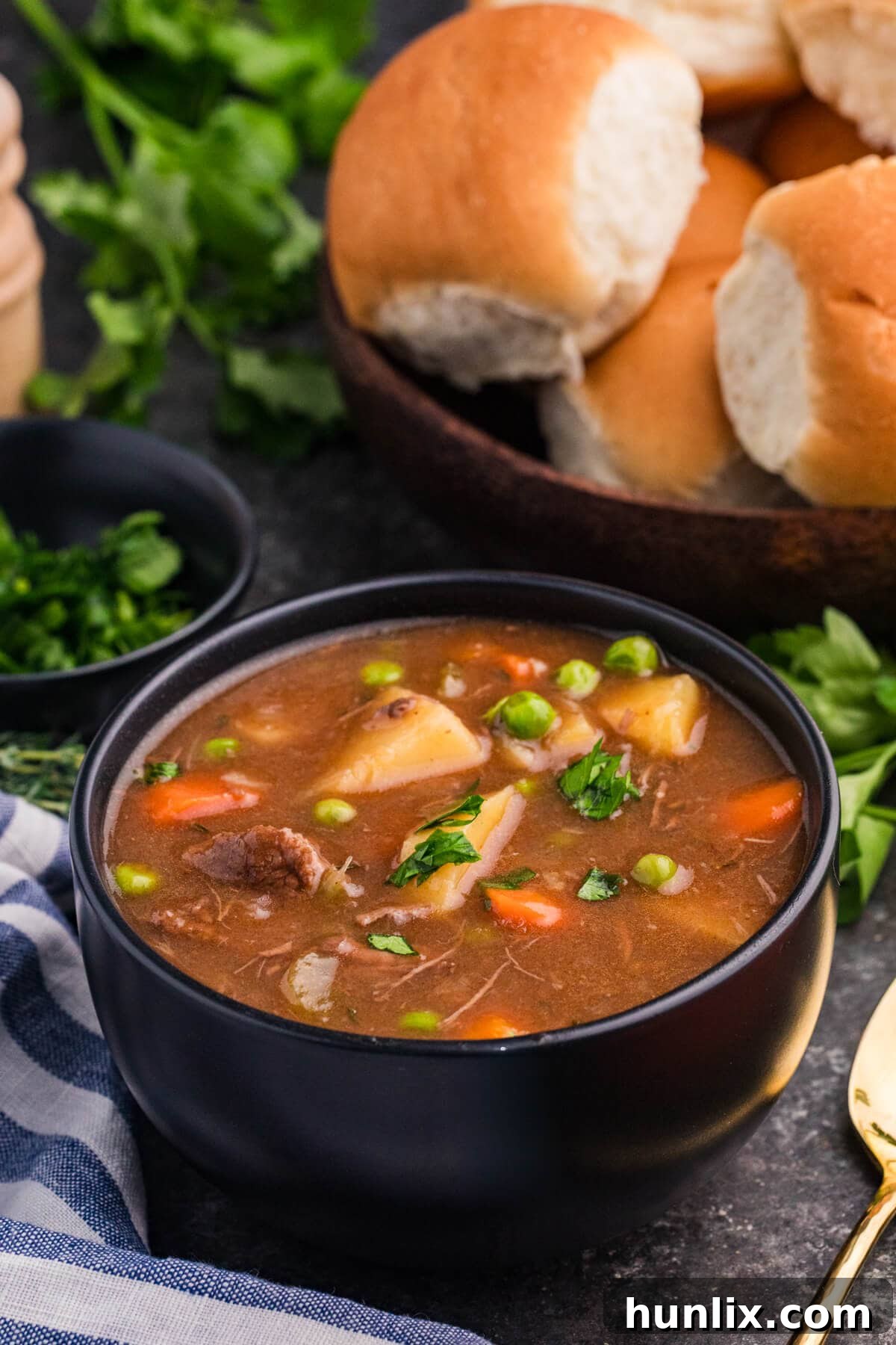 A comforting bowl of old fashioned beef stew, brimming with tender meat, chunky potatoes, carrots, and vibrant peas, garnished with fresh herbs and served alongside crusty dinner rolls on a wooden surface.