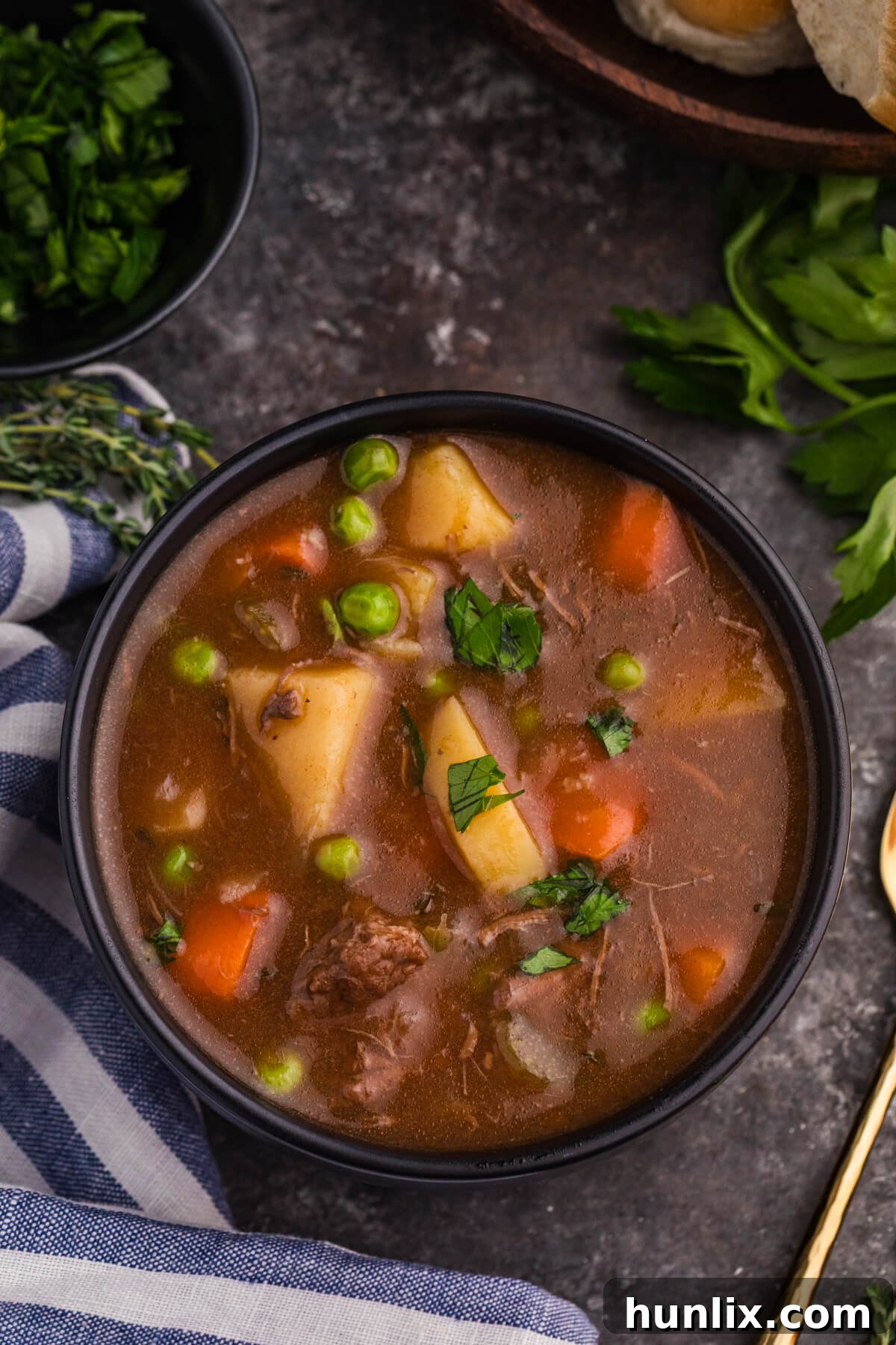 A top-down view of a rustic black bowl filled with hearty beef stew, showcasing tender shredded beef, chunky carrots, potatoes, and bright green peas in a rich, dark broth, garnished with fresh parsley.