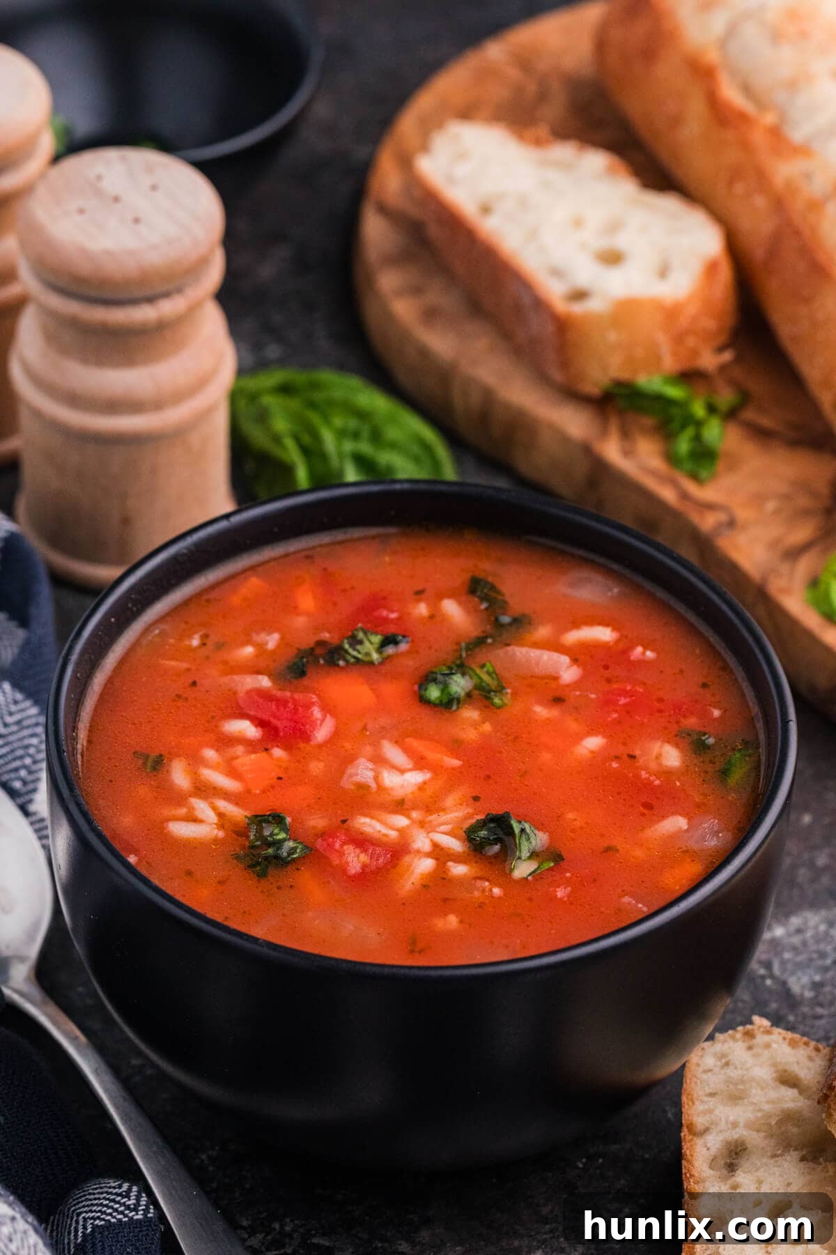 A black bowl brimming with rustic tomato rice soup, garnished with fresh green herbs and served alongside slices of crusty artisan bread, ready to be enjoyed.