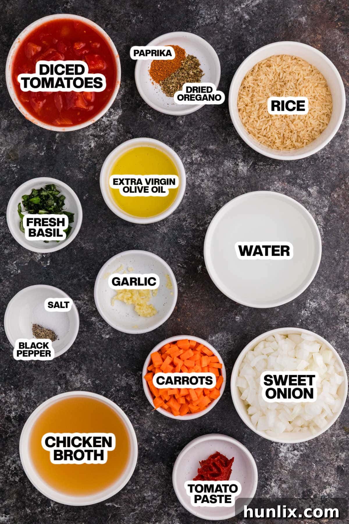 A collection of fresh, vibrant ingredients for tomato rice soup laid out on a dark background, including olive oil, onions, carrots, garlic, spices, canned tomatoes, broth, rice, and fresh basil.