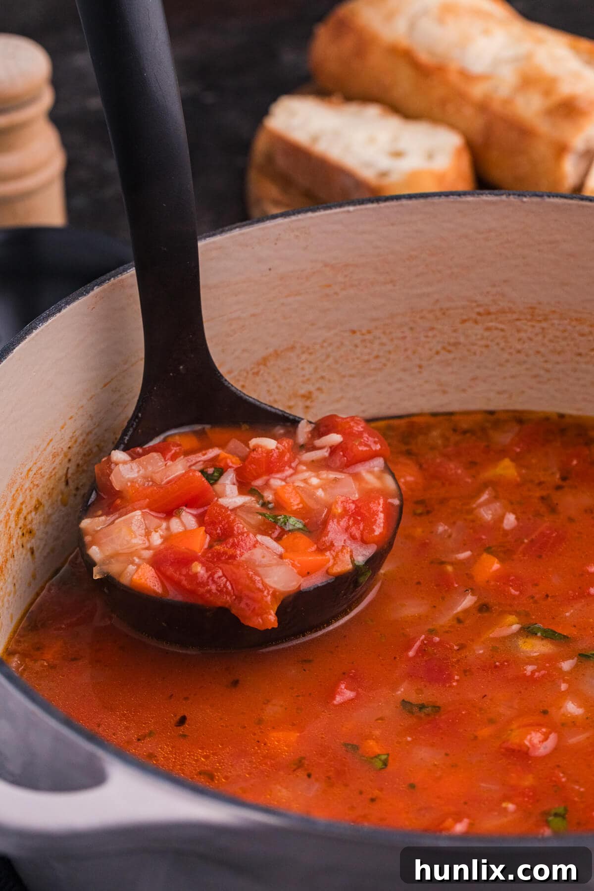 A ladle serving a generous portion of thick, red tomato rice soup, filled with visible diced tomatoes, carrots, and onions, from a large Dutch oven, garnished with fresh herbs.
