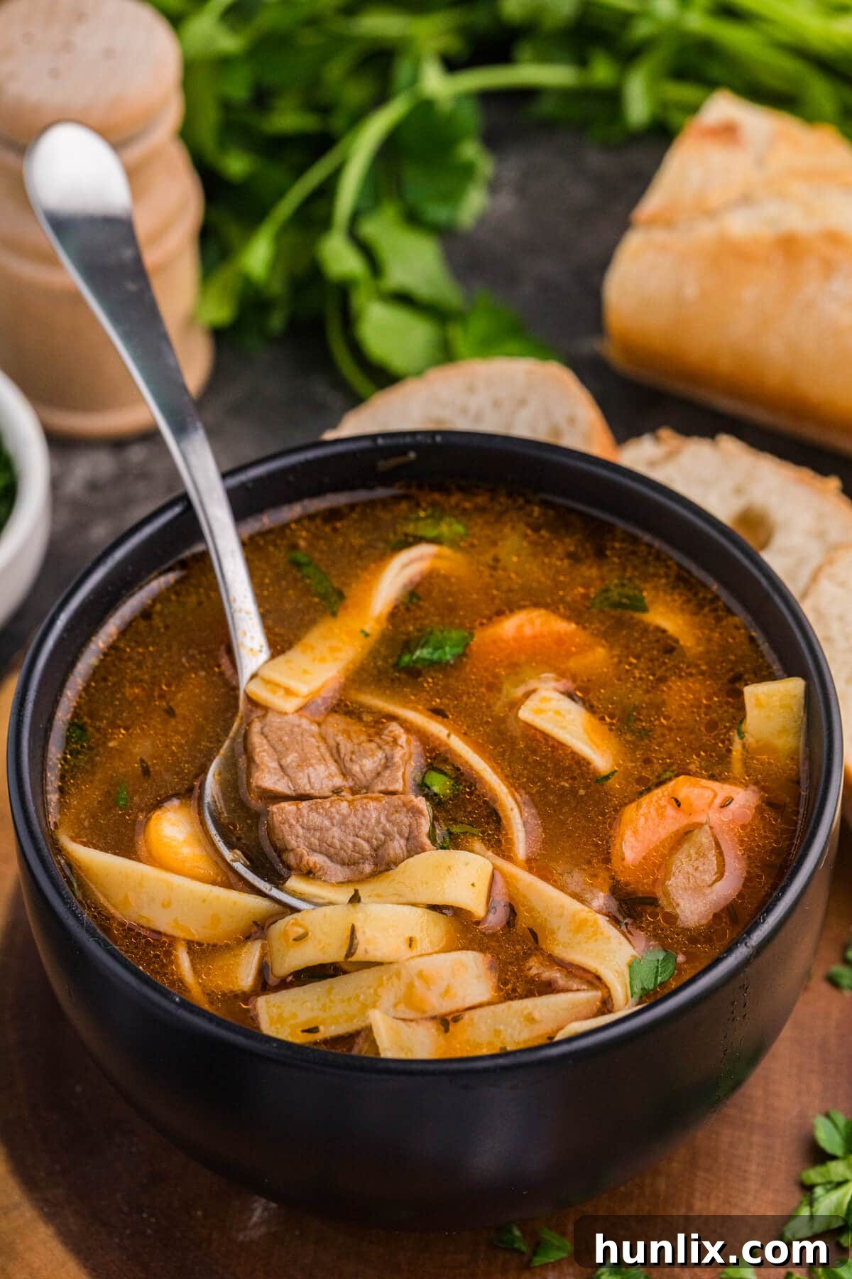 Spoonful of beef noodle soup with carrots and noodles in a black bowl, fresh bread in background.