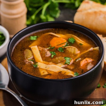 Bowl of beef and noodle soup with vegetables, garnished with parsley, served with bread.