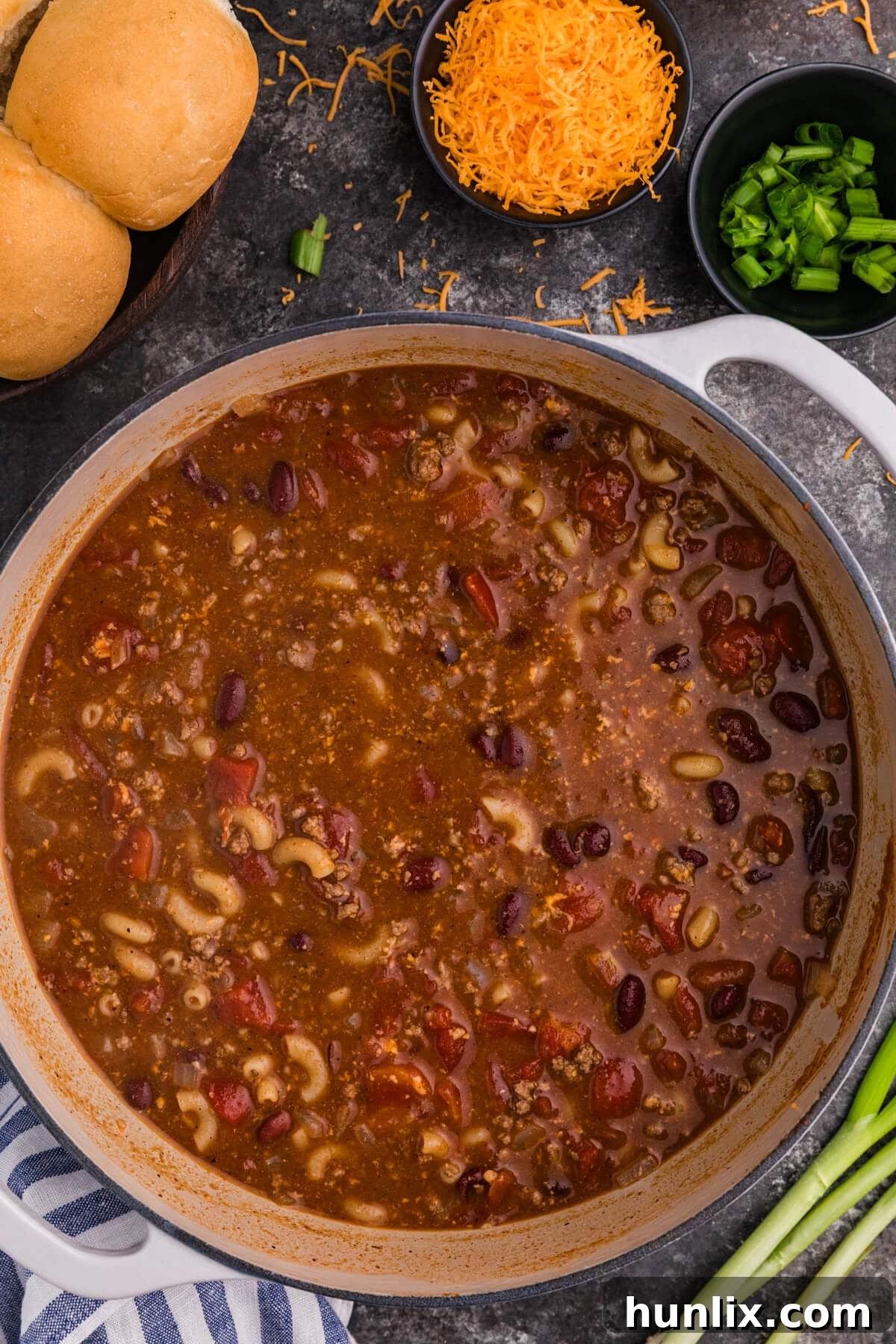 Chili Mac Soup in a white Dutch oven with elbow macaroni, ground beef, kidney beans, and tomatoes, served with shredded cheddar, green onions, and dinner rolls.