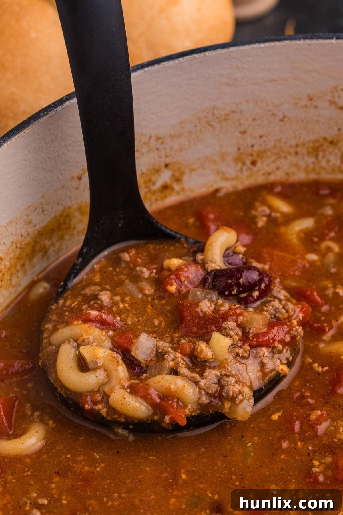 Close-up of a ladle scooping hearty macaroni, ground beef, kidney beans, and tomato broth from a white Dutch oven.