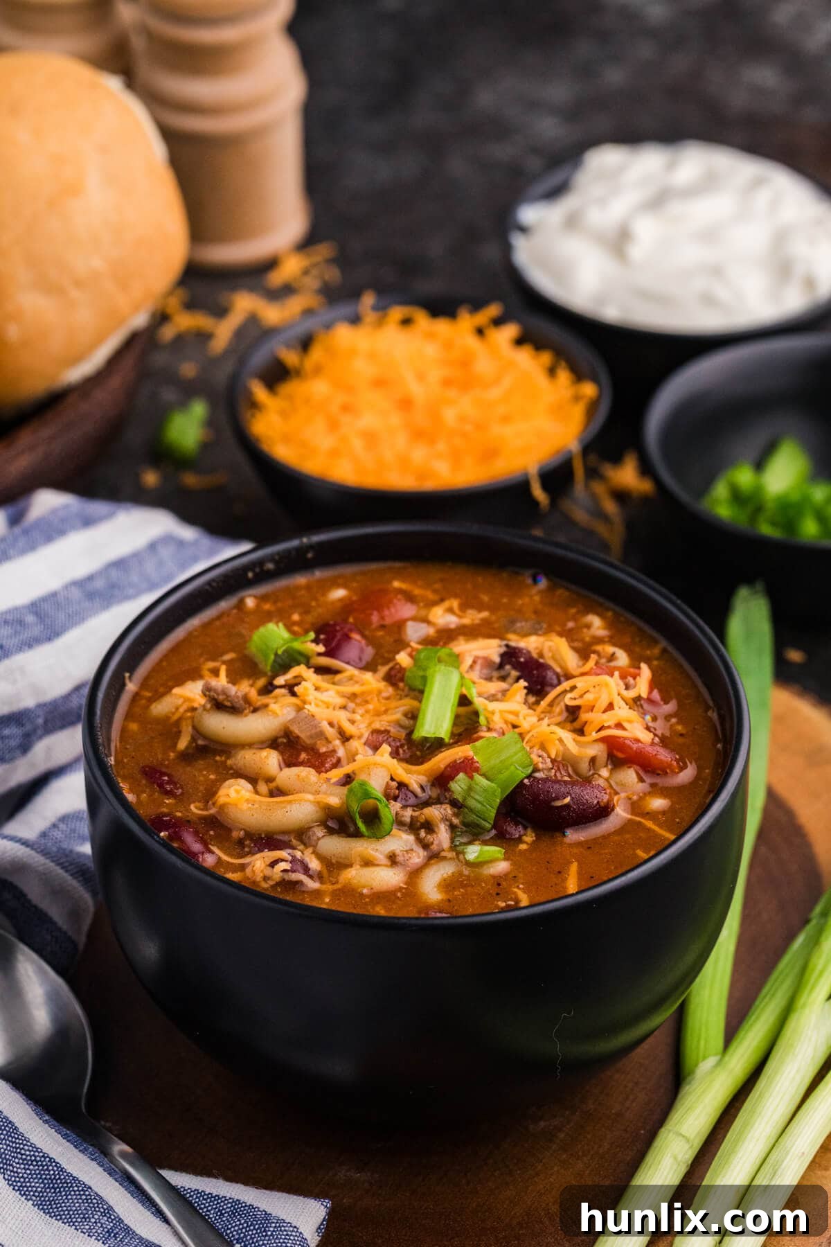 Black bowl of macaroni, ground beef, kidney beans, and tomato soup topped with shredded cheddar and green onions, with rolls and sour cream in the background.