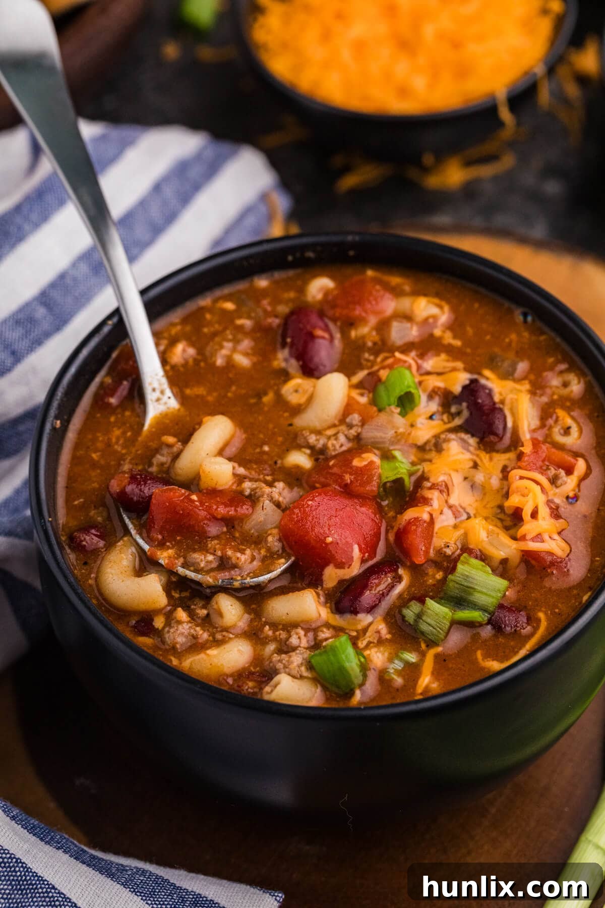 Spoon lifting macaroni, ground beef, kidney beans, tomatoes, and melted cheddar from a black bowl of savory tomato-based soup.
