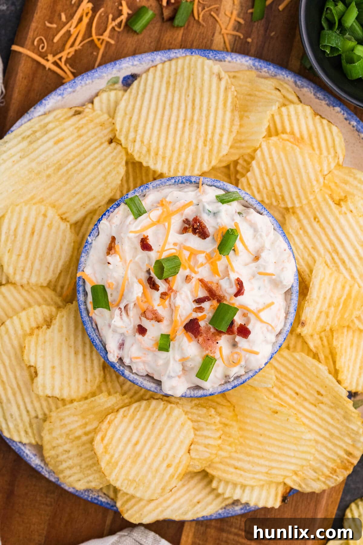 Absolutely Irresistible Dip 2 Crack Dip topped with crispy bacon, sharp cheddar cheese, and vibrant green onions, served alongside ridged potato chips on a rustic wooden board, ready for a party.