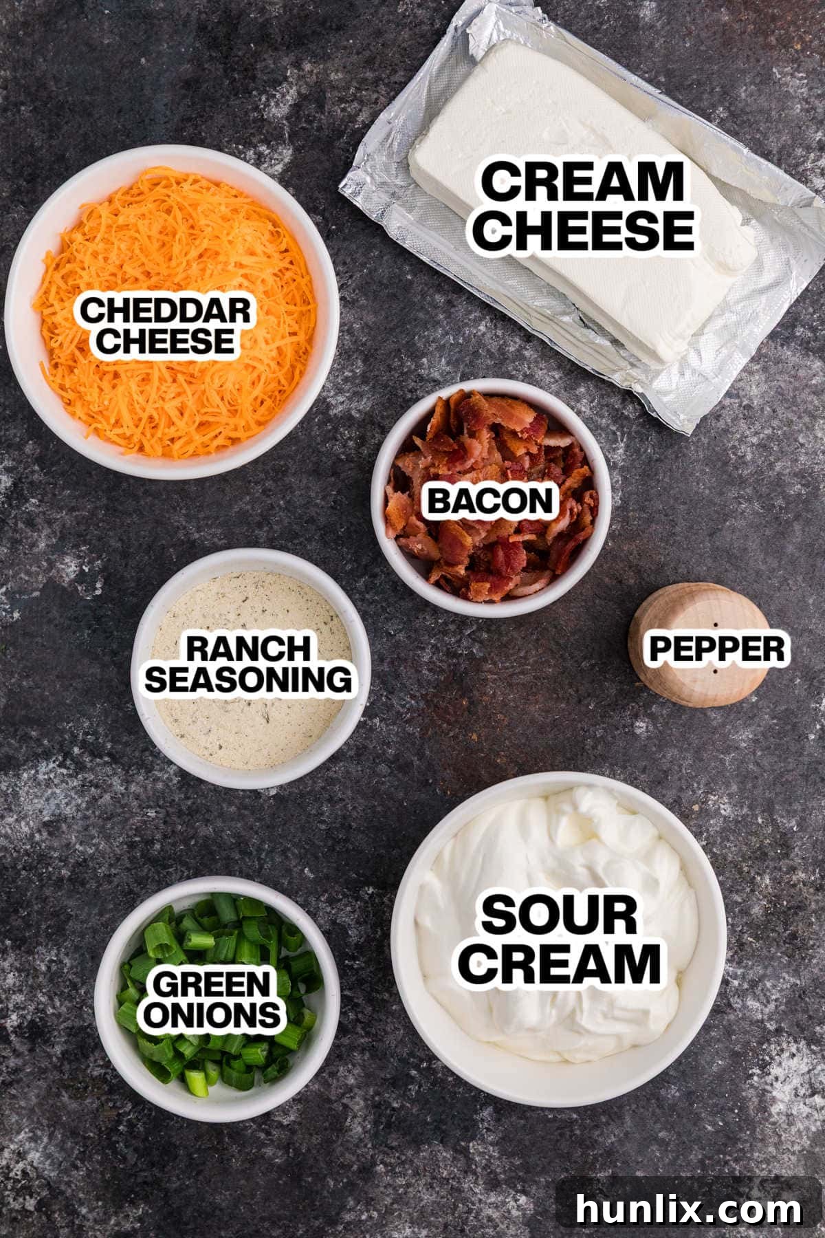 Absolutely Irresistible Dip 3 Assortment of fresh ingredients for Crack Dip laid out on a dark background, including cream cheese, sour cream, cheddar, bacon, ranch seasoning, and green onions.