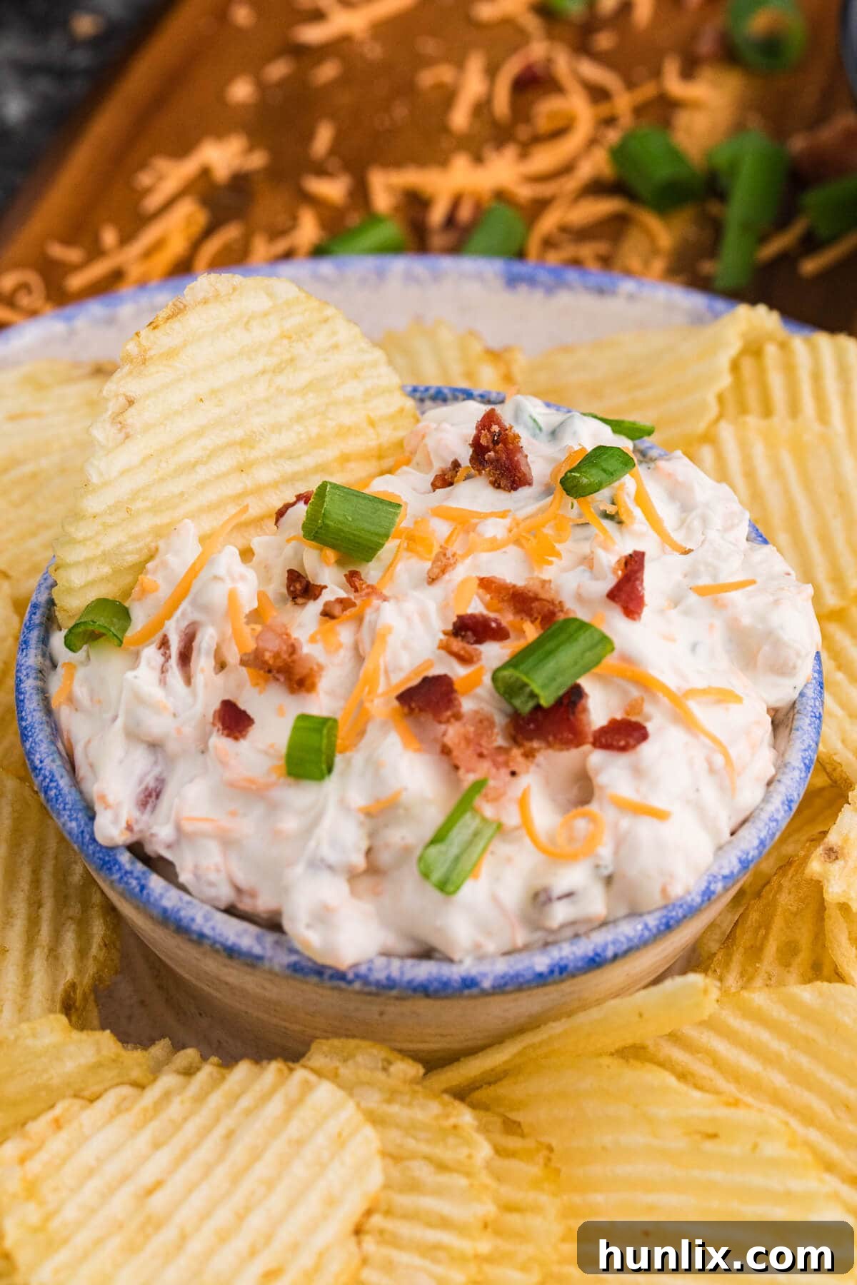 Absolutely Irresistible Dip 6 A tantalizing close-up shot of loaded bacon cheddar ranch dip with visible bits of crispy bacon and vibrant green onions, accompanied by crunchy potato chips.