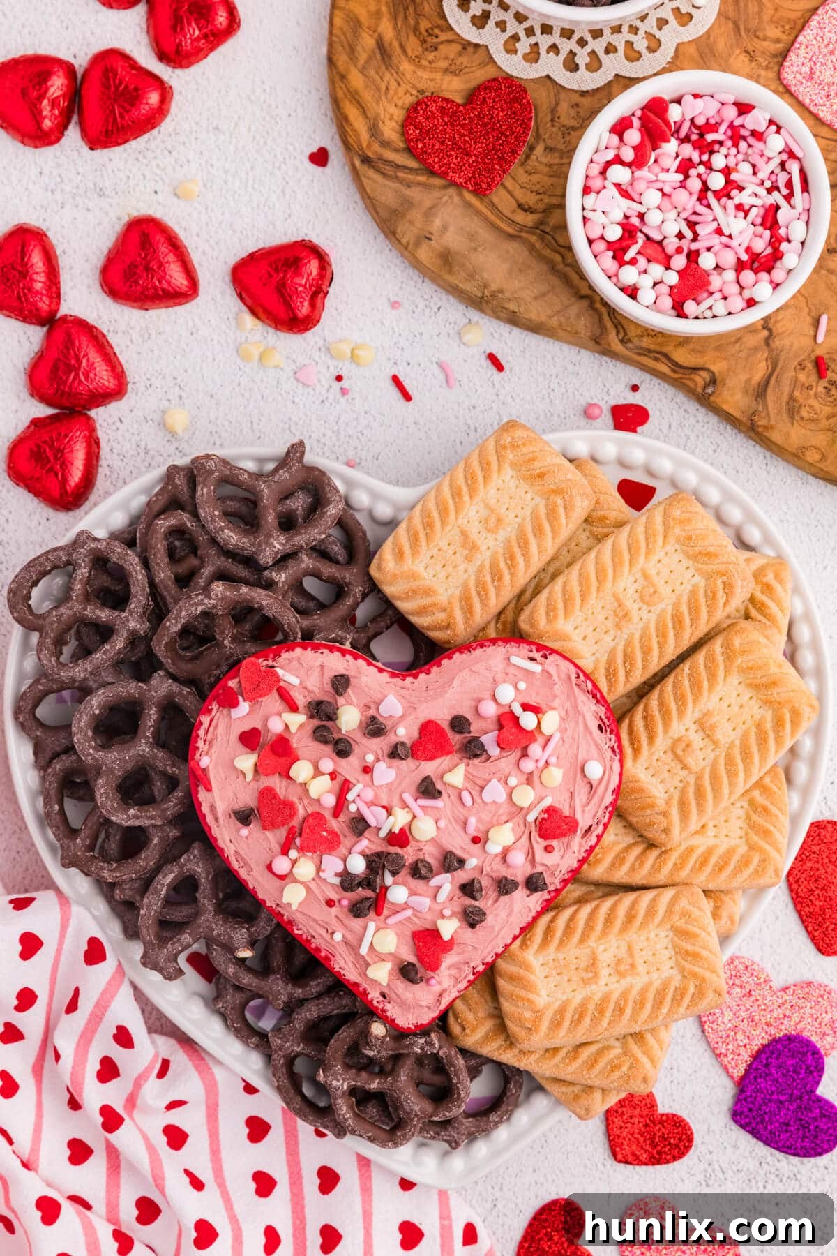 Red Velvet Cheesecake Dip in a heart-shaped bowl topped with Valentine’s sprinkles, surrounded by chocolate pretzels, butter cookies, and red foil-wrapped chocolate hearts.