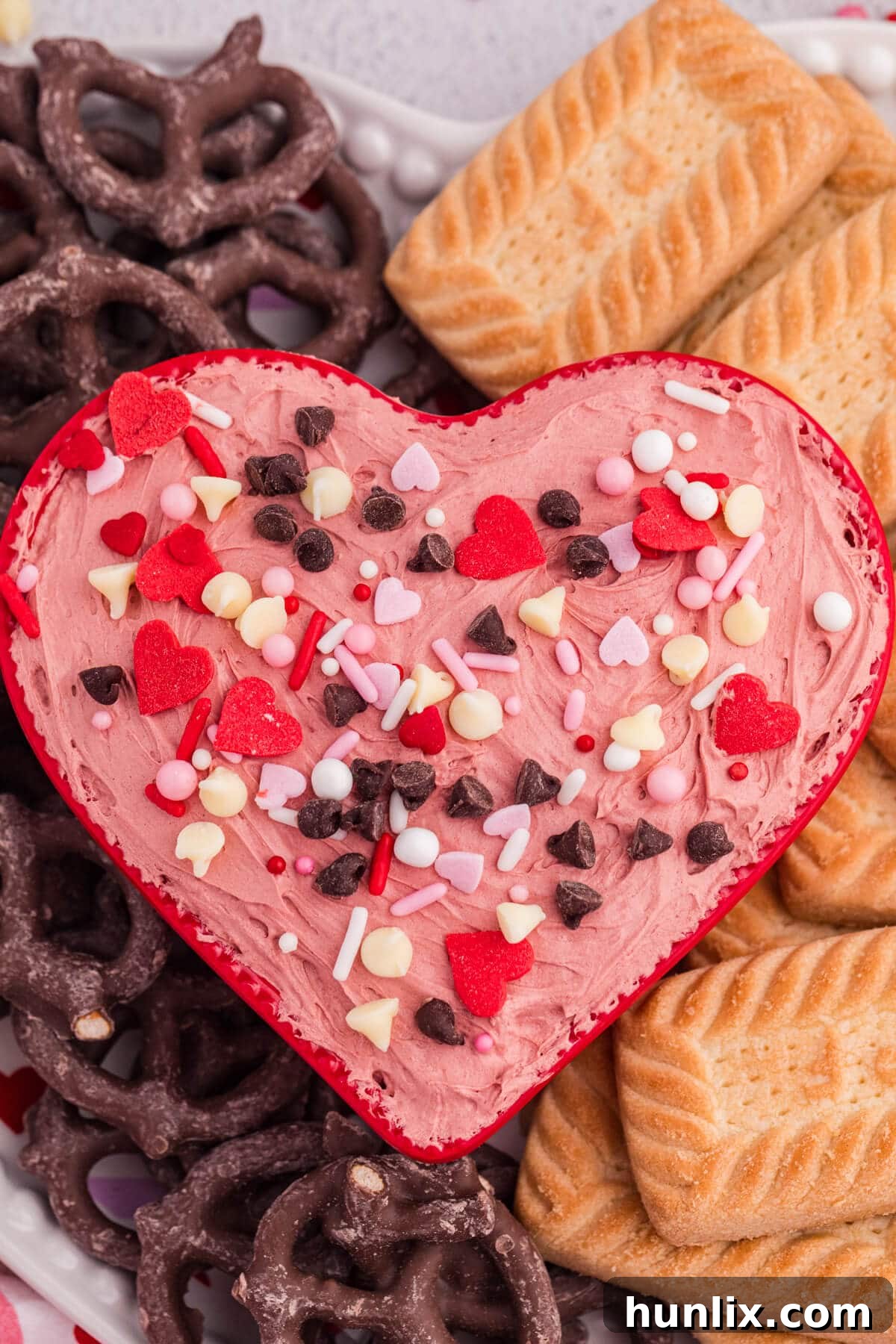 Close-up of pink cheesecake dip in a heart-shaped dish with candy hearts and chocolate chips, served with chocolate-covered pretzels and cookies.